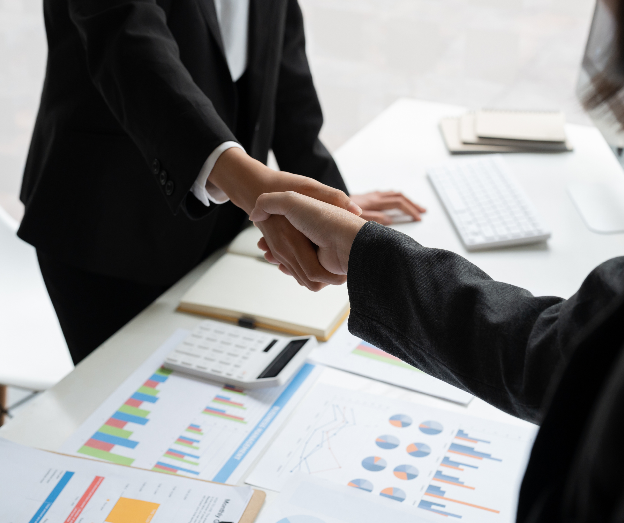 Two business professionals in suits shaking hands over a desk with financial charts, calculator, notebook, keyboard, and documents.
