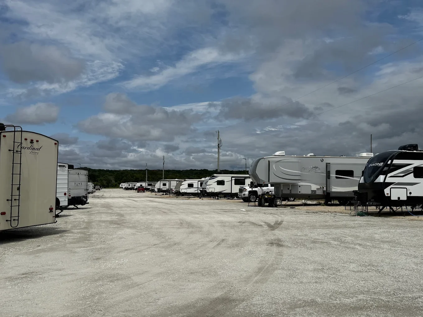 A gravel lot filled with recreational vehicles, including travel trailers and motorhomes, under a partly cloudy sky with scattered clouds and some green trees in the distance.