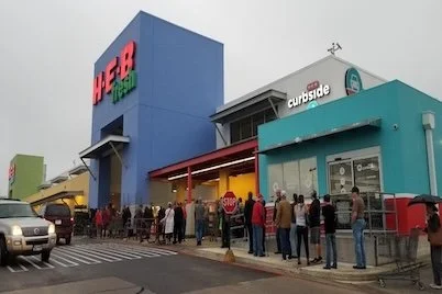 People are standing in line outside a colorful H-E-B grocery store with several other storefronts nearby.