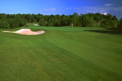 A golf course with a sand trap, green grass, and a flag on the hole, surrounded by trees under a blue sky.