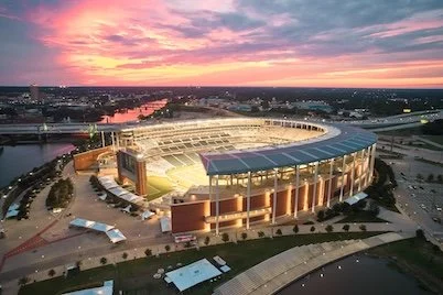 A large sports stadium during sunset with a brightly lit exterior and surrounding roads, overlooking a cityscape with a river.