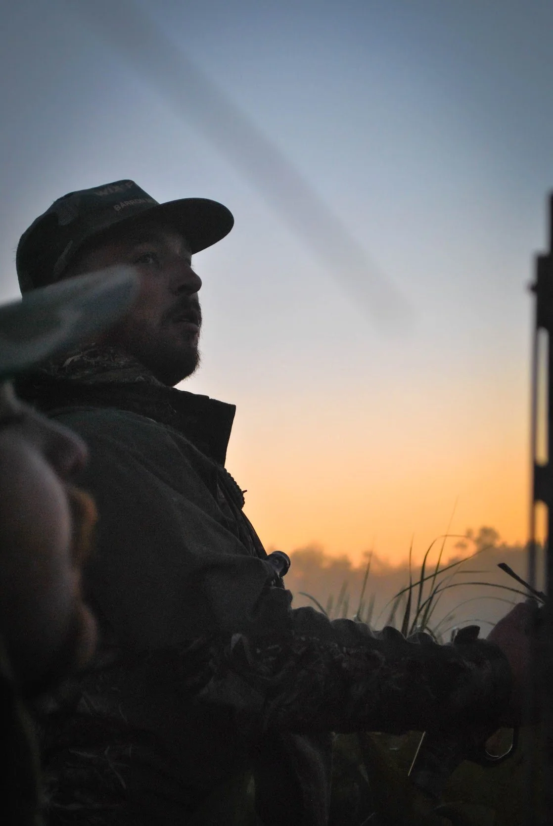 A man in hunting gear with a cap, sitting outdoors at sunrise, possibly operating a machine or weapon, with grass in the foreground and a orange and blue sky in the background.