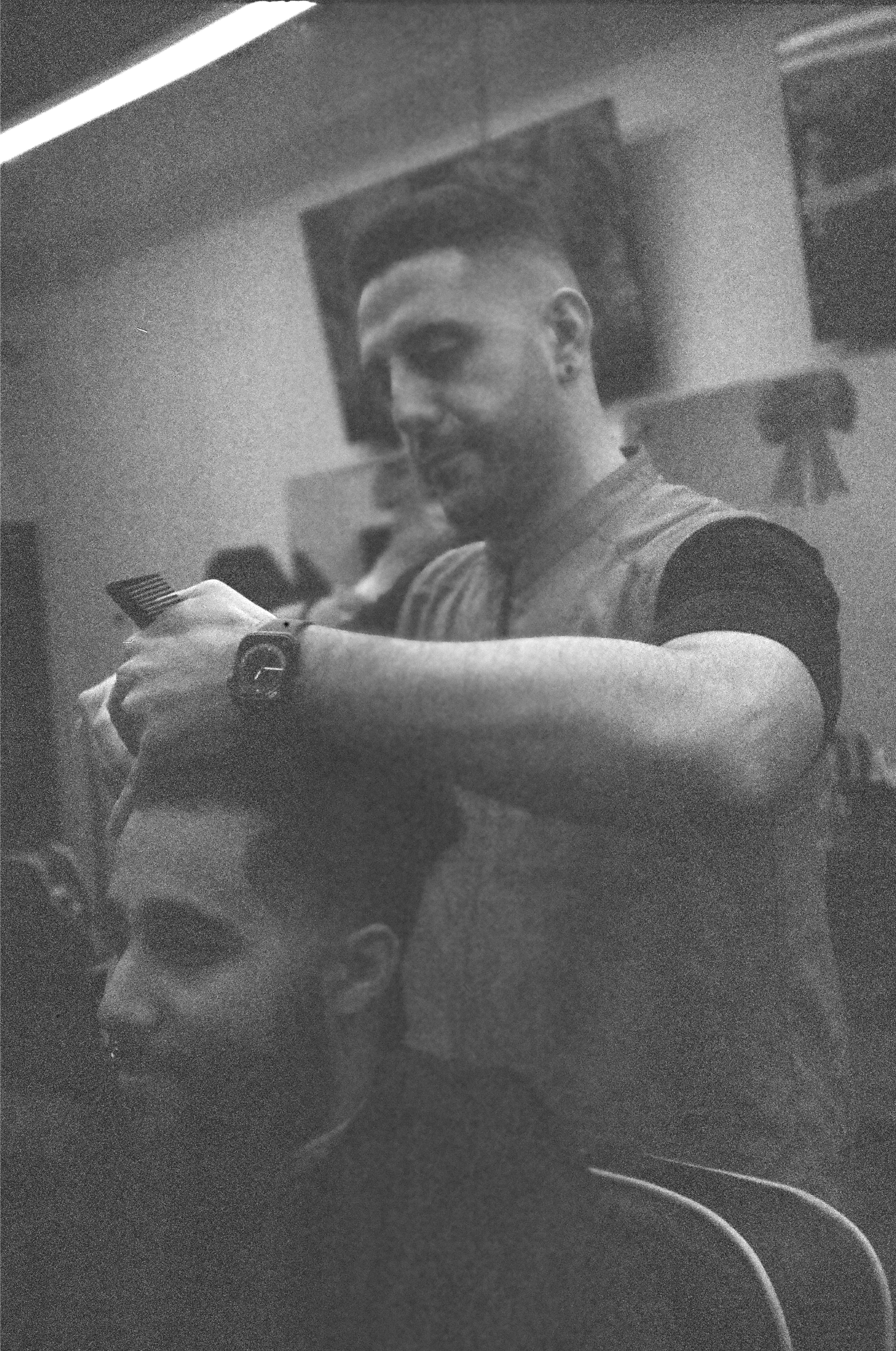 Black and white photo of a man with short hair, smiling, making a peace sign with his hand, in a room with a large screen and other people in the background.