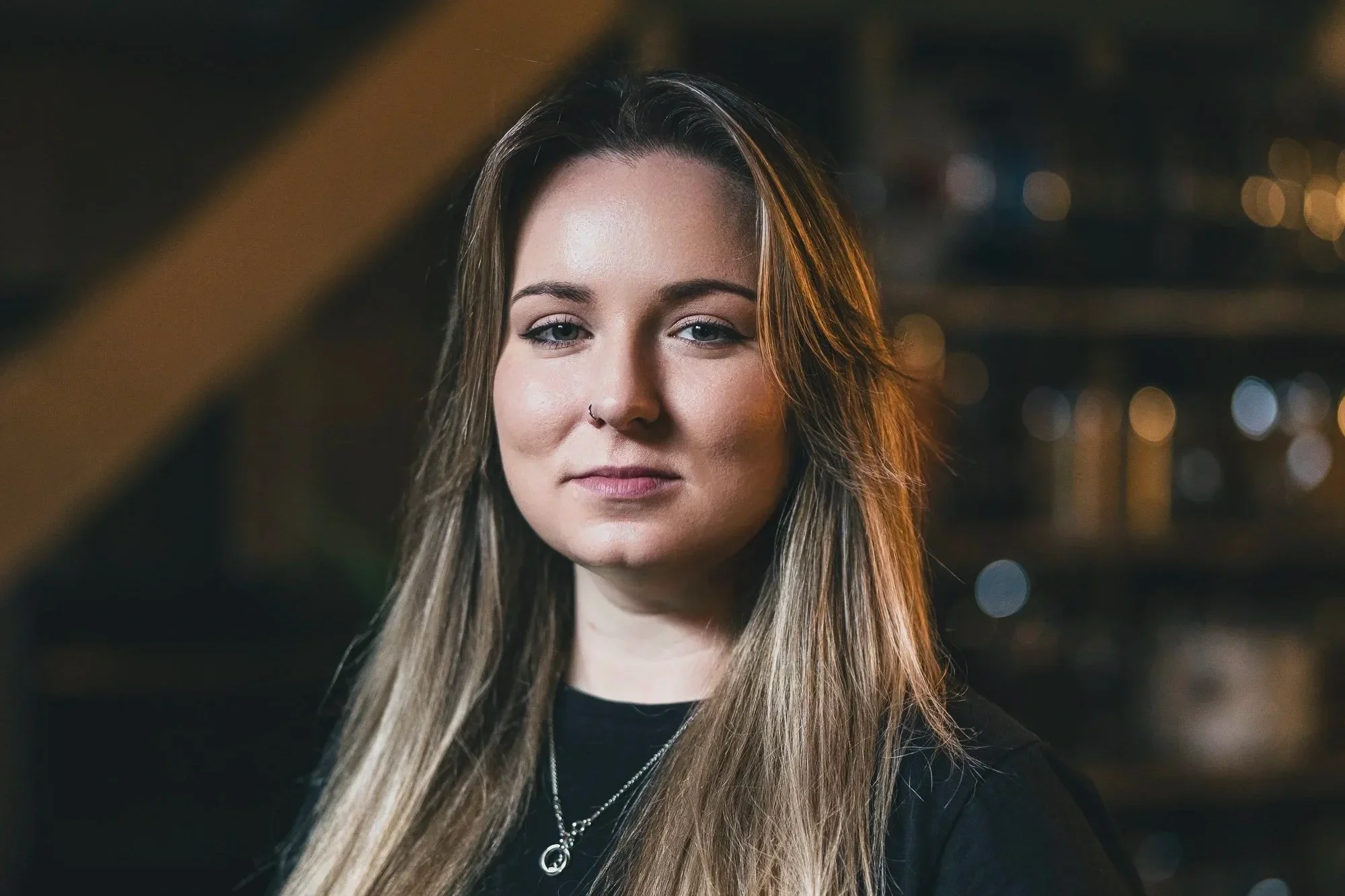 A young woman with long, light brown hair and a nose piercing, wearing necklaces, looks confidently at the camera in a dimly lit indoor setting.