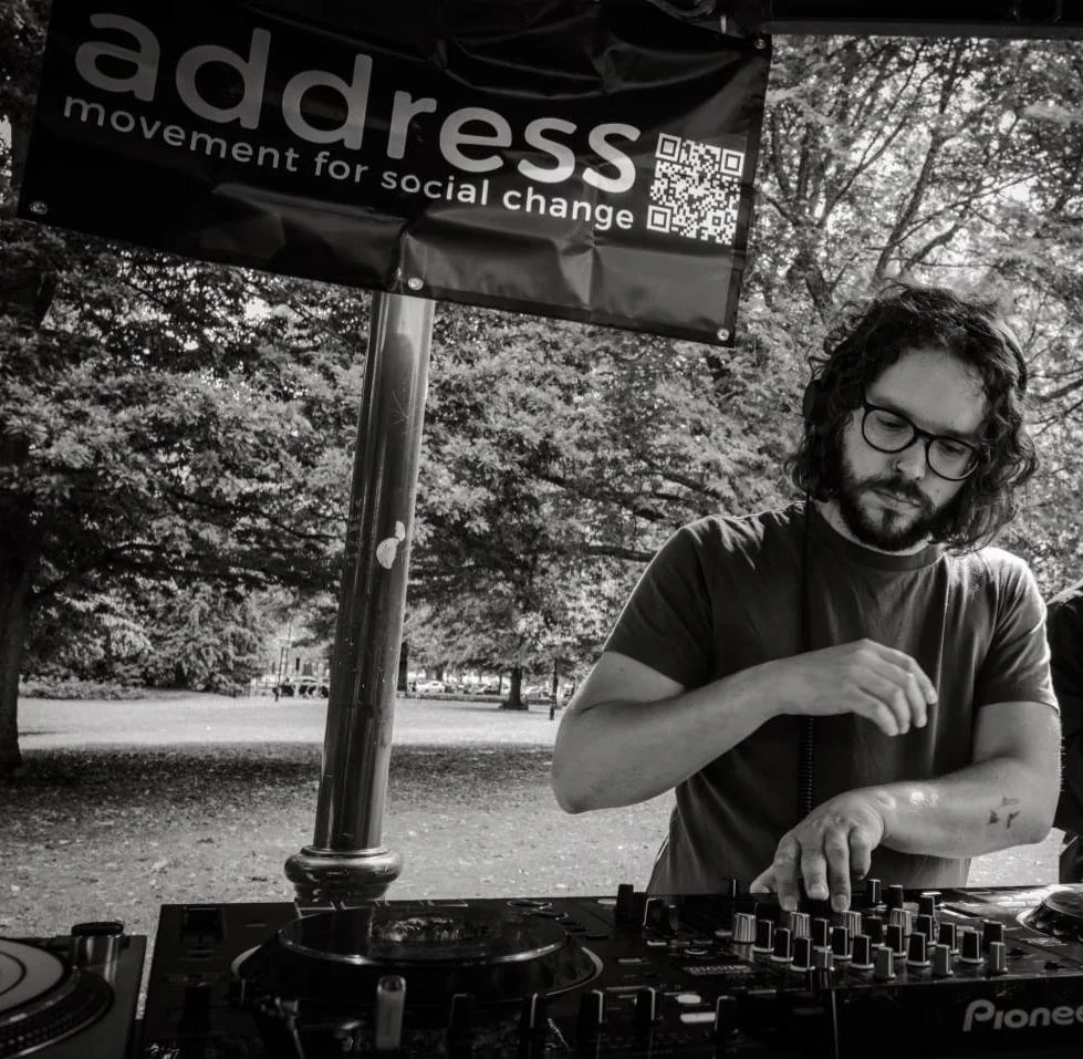 DJ with curly hair, glasses, and headphones, working on a DJ console with colorful lights.