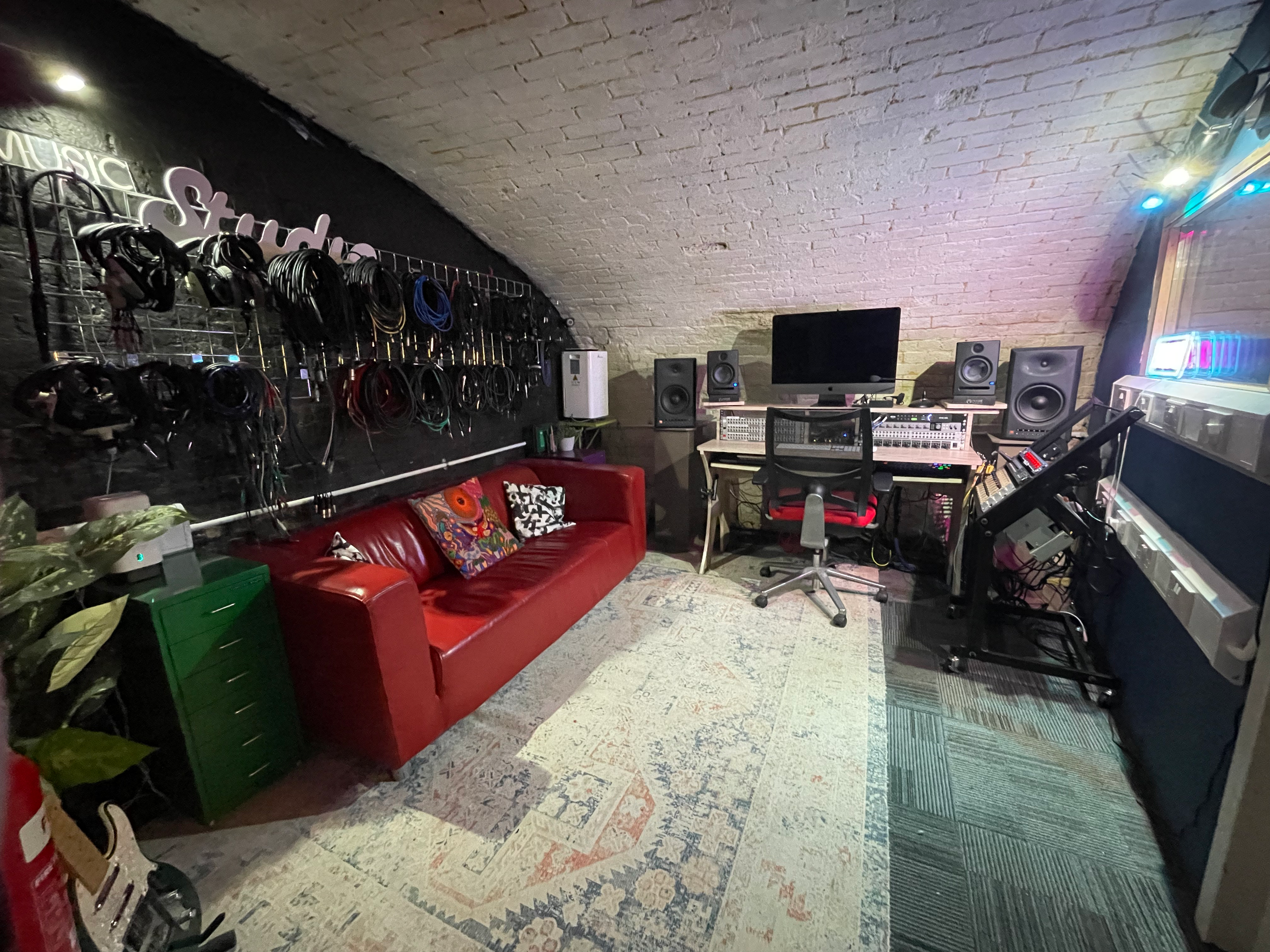A music studio with a red leather couch, patterned pillows, a green side table, and a large mixing console with multiple speakers and monitors. The room has a curved brick ceiling and walls with audio equipment, cables, and a computer setup.