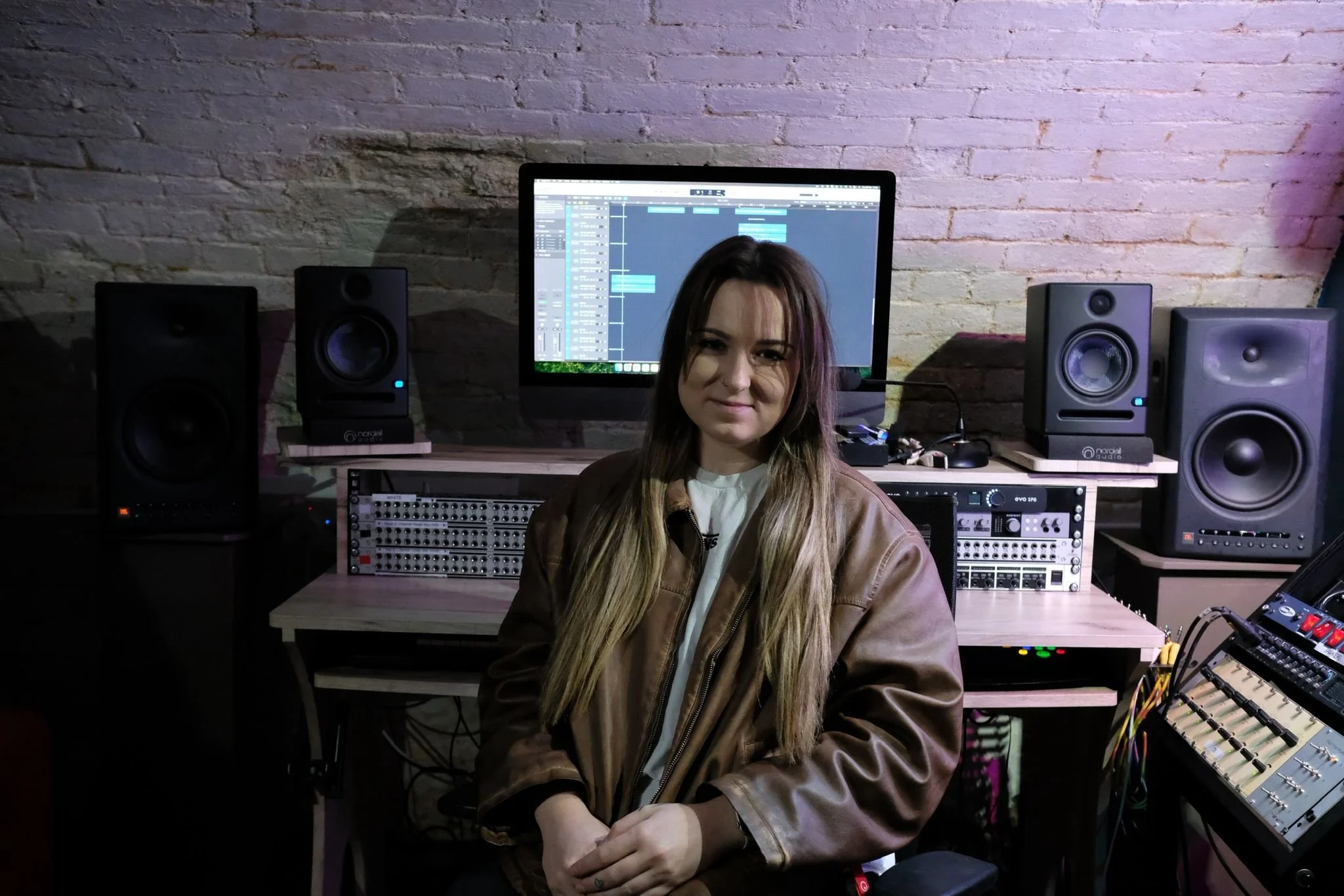 A woman with long hair and a brown leather jacket sitting in a music studio in front of a computer monitor with a digital audio workstation open, surrounded by studio speakers, audio equipment, and a modular synthesizer.