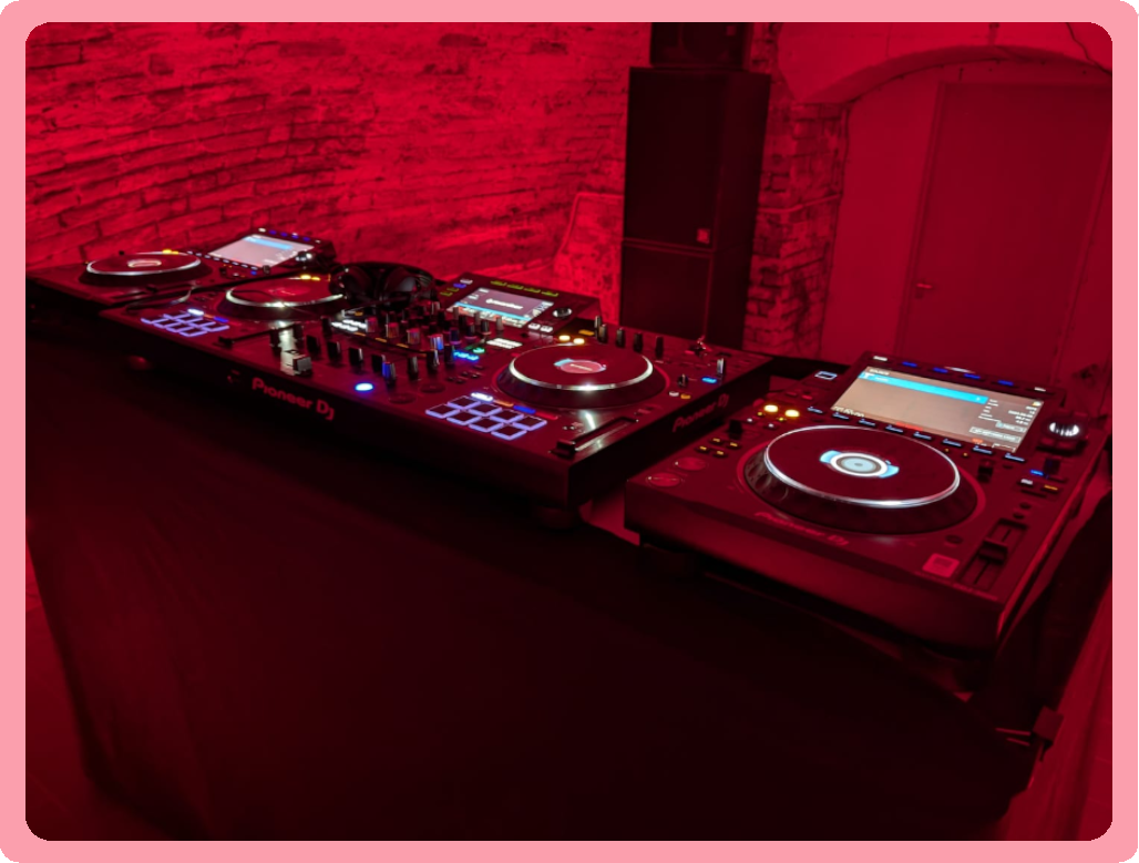 A DJ setup with two Pioneer DJ controllers and a mixer on a table, illuminated by red lighting, against a brick wall background.