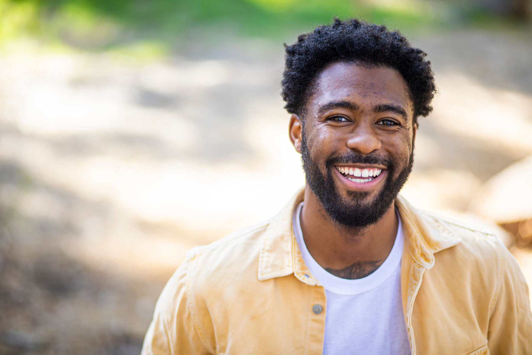 A muslim smiling man with a beard and curly hair outdoors, wearing a yellow jacket over a white shirt after receiving therapy with a salamah counsellor.