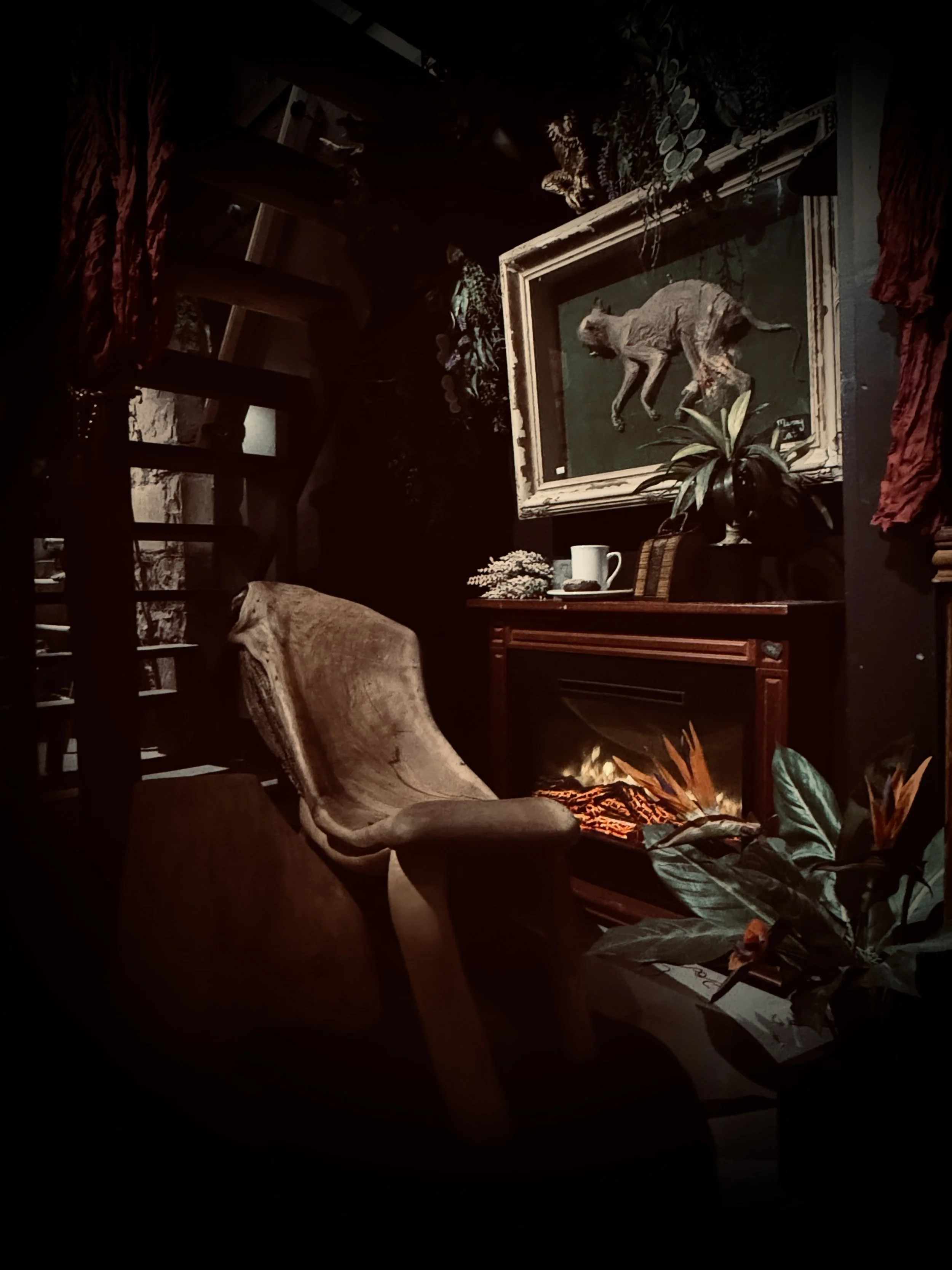 A cozy room with a wooden chair, a framed artwork of a cat, a small table with a mug, plants, and a view of a fireplace.