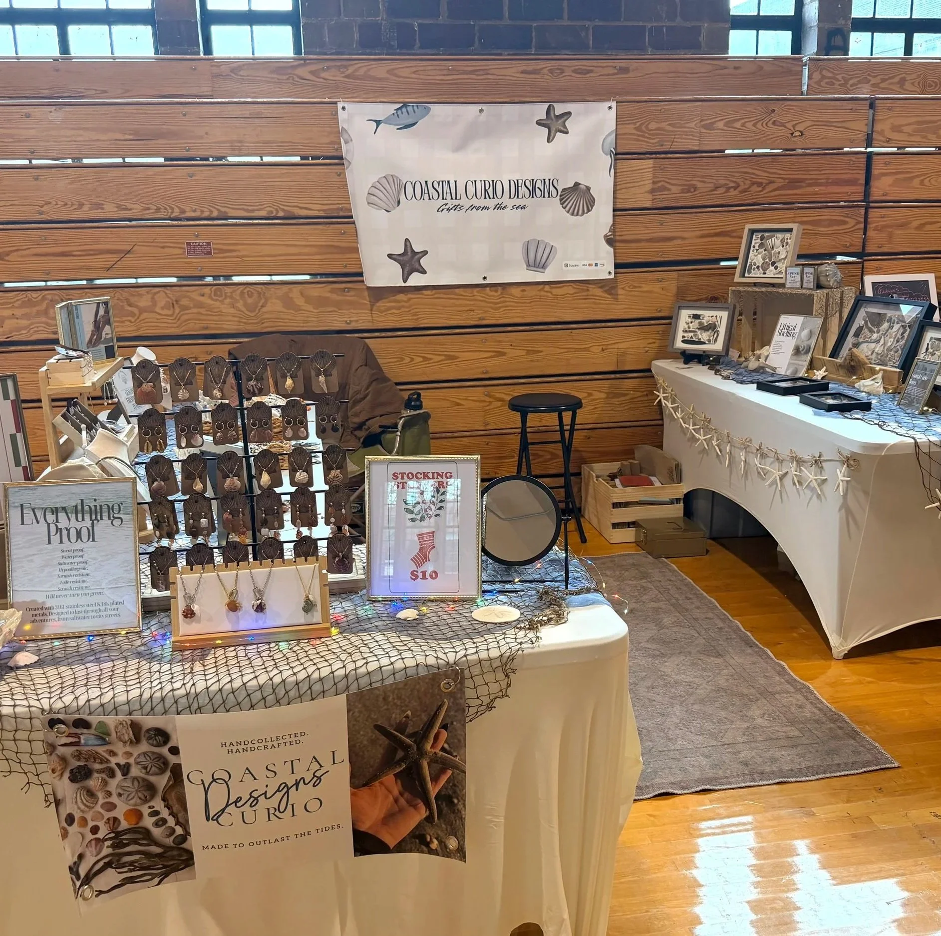 Display booth with jewelry and coastal-themed decor, featuring necklaces, earrings, and framed artwork, in a room with wooden walls and hardwood floors.