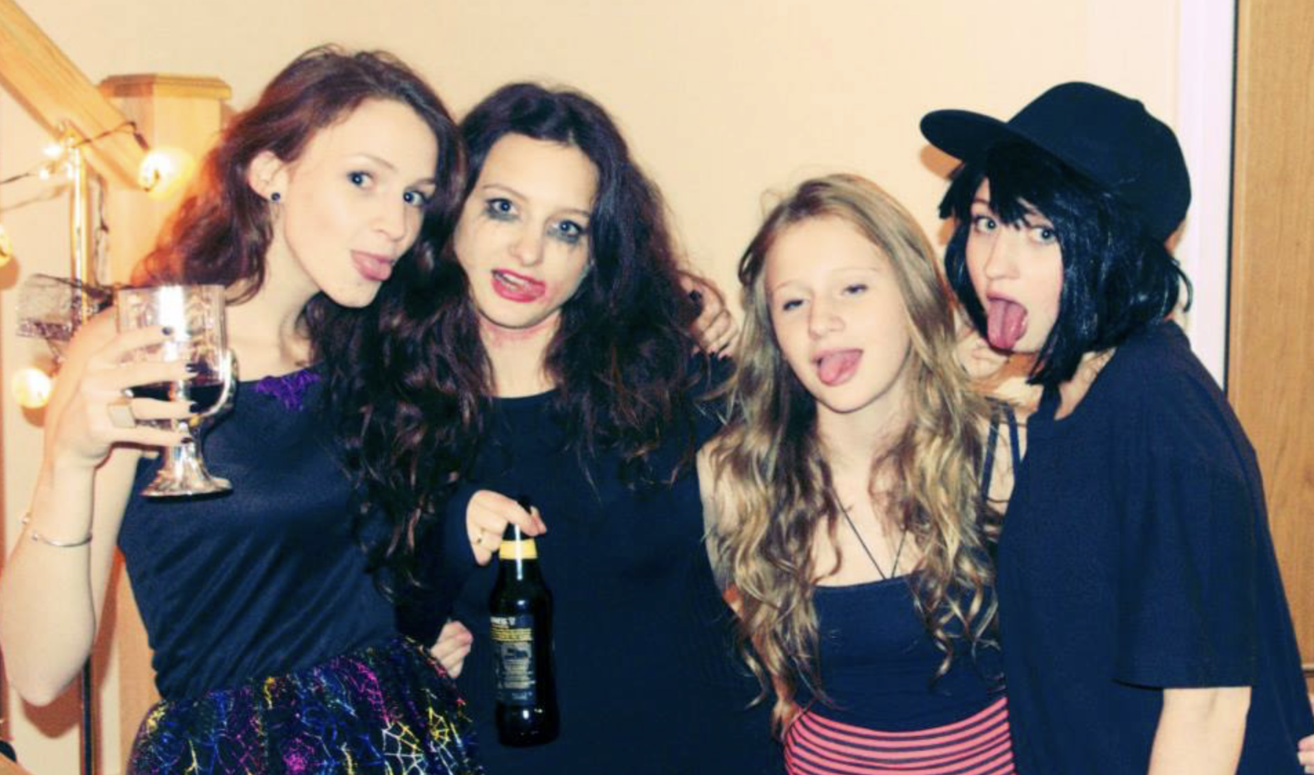Four young women at a party making funny faces, with two sticking out their tongues and two with their eyes closed, in front of a light-colored wall with a wooden door.