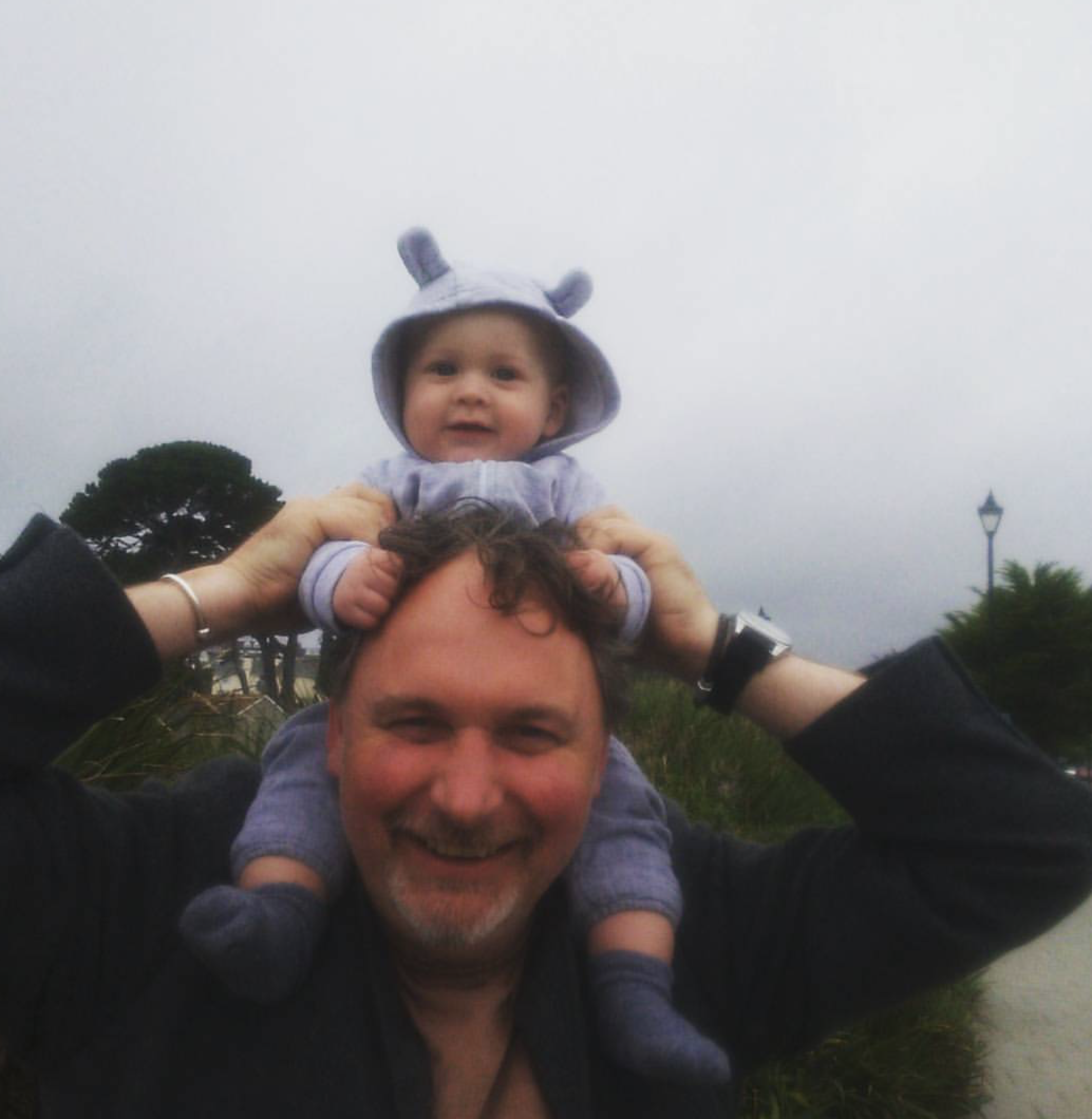 A man smiling while carrying a young child on his shoulders outdoors on a cloudy day.