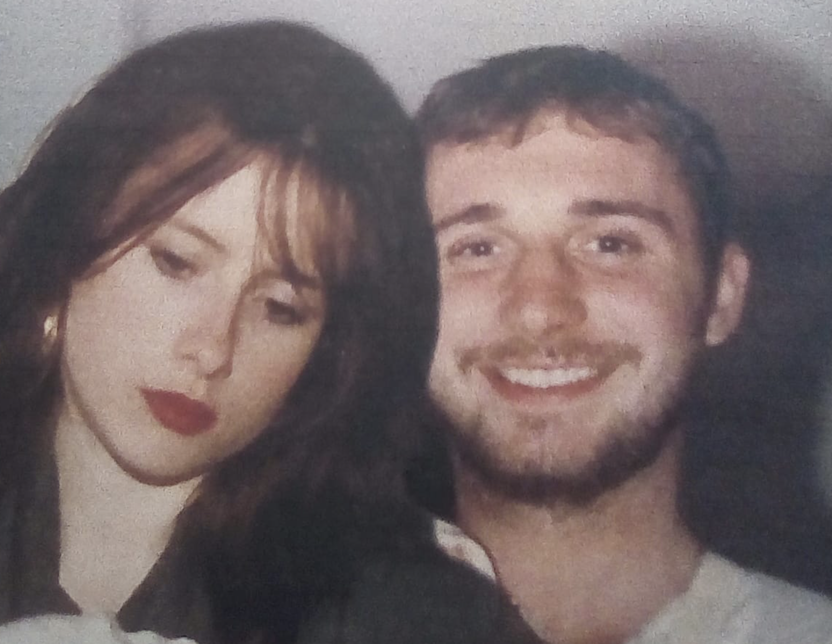 A woman with dark brown hair and red lipstick and a man with short dark hair and a beard smiling, close together in a photo.