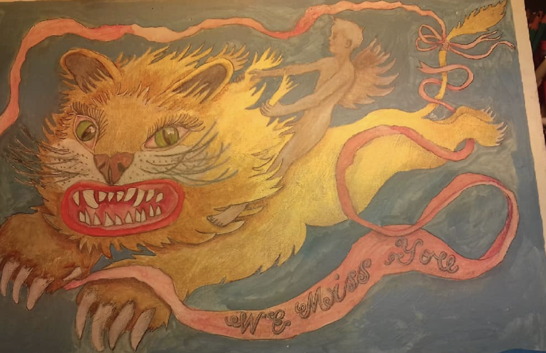 A painting of a fierce lion with green eyes, sharp teeth, and a flowing yellow mane. A small winged fairy or angel figure is located above the lion with wings spread. A pink ribbon winds around the lion with the words 'We Move As One' written on it.