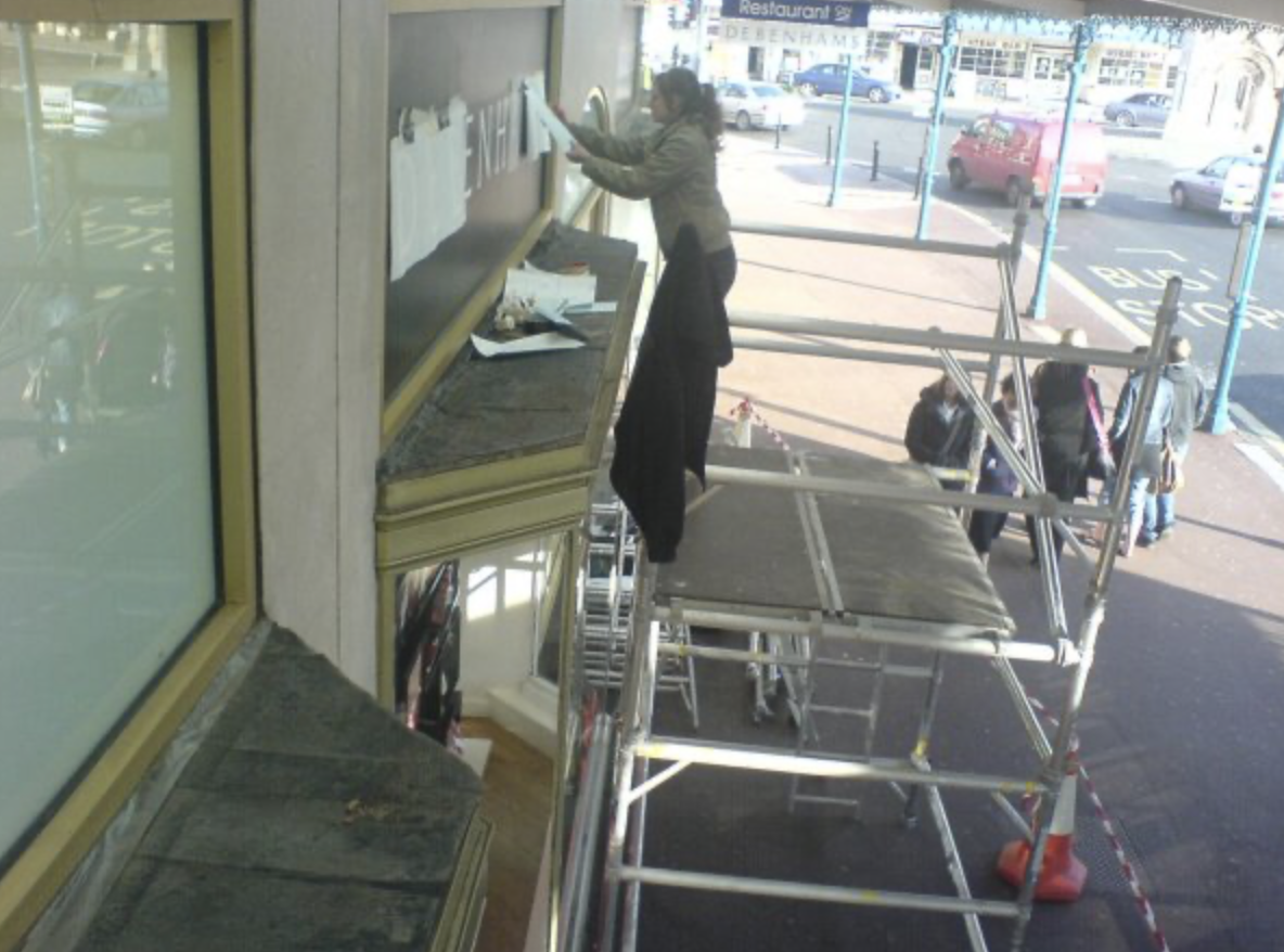 A woman on scaffolding outside a building is putting up papers or signs in a display case. Several people are walking on the sidewalk in the background.