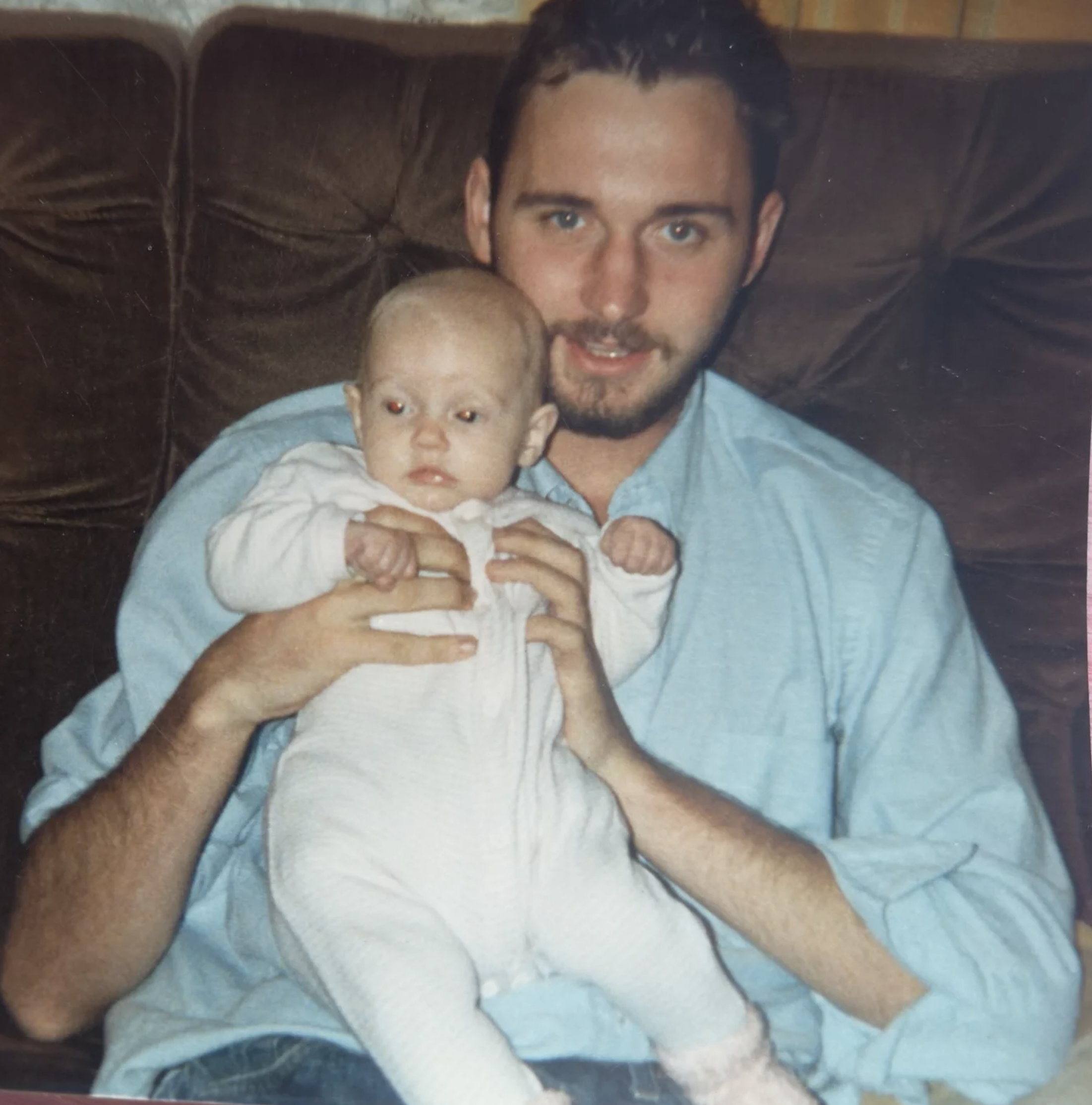 A man with a beard holding a baby with blond hair and blue eyes, sitting on a dark brown couch.