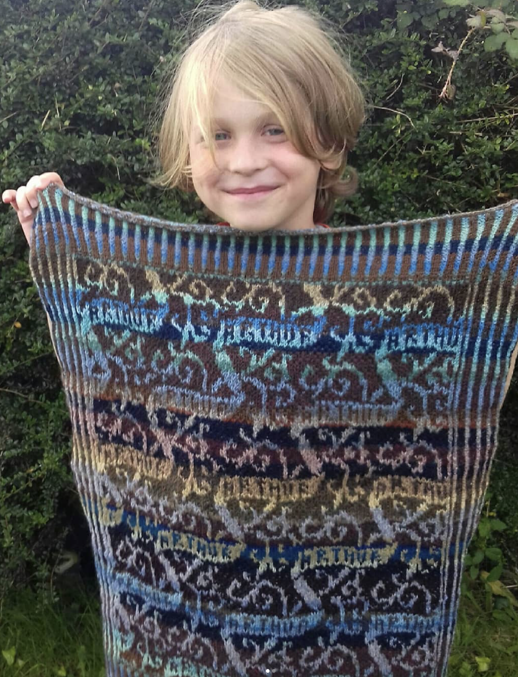 A young boy with blond hair smiling while holding a colorful, woven textile or blanket outdoors in front of greenery.