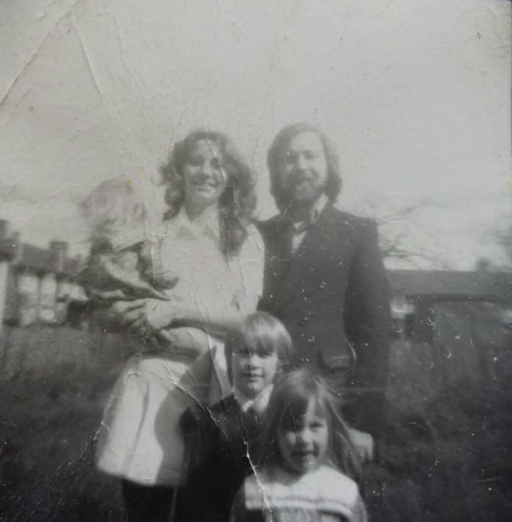 A black and white photo of two adults and two children outdoors, with a building and trees in the background.