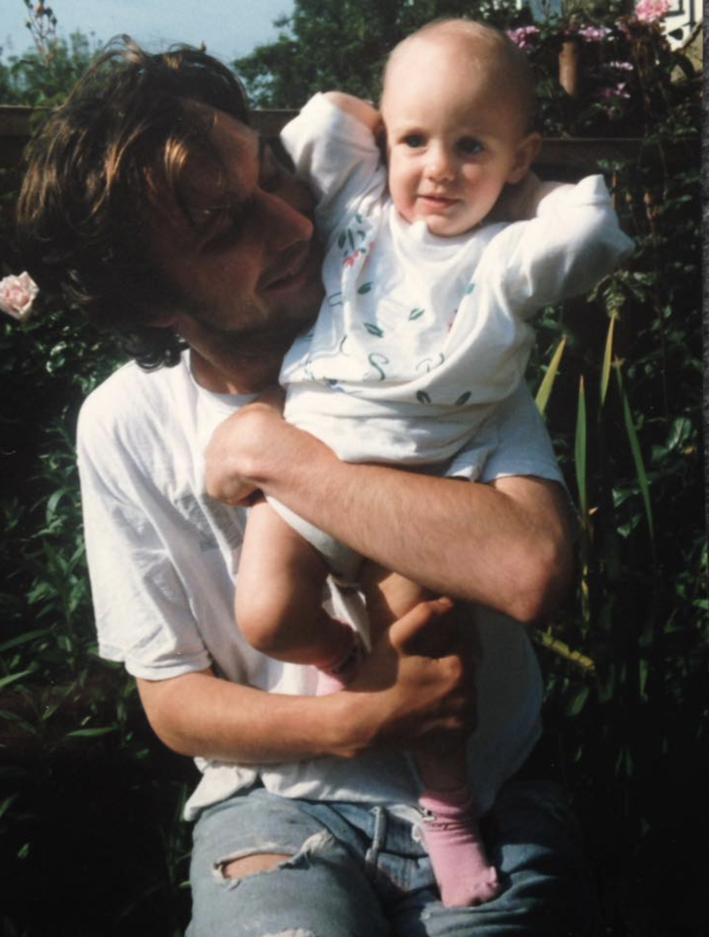 A man with curly hair and glasses holding a young child in his arms outdoors among green plants and flowers.