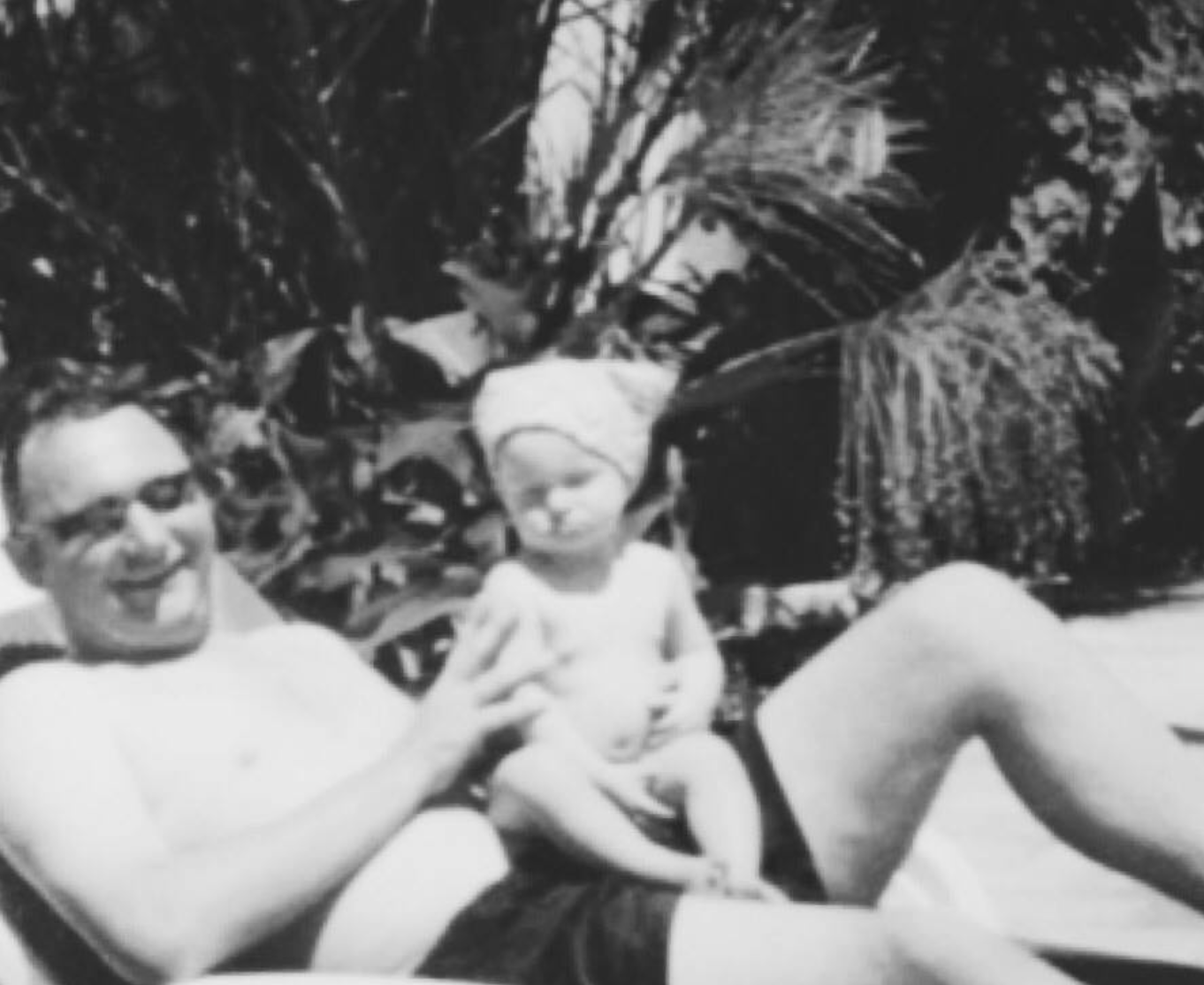 Black and white photo of a man with glasses and a young girl sitting outdoors among large plants. The man is lying back with a big smile, and the girl is smiling while sitting on his lap, with a paper hat on her head.