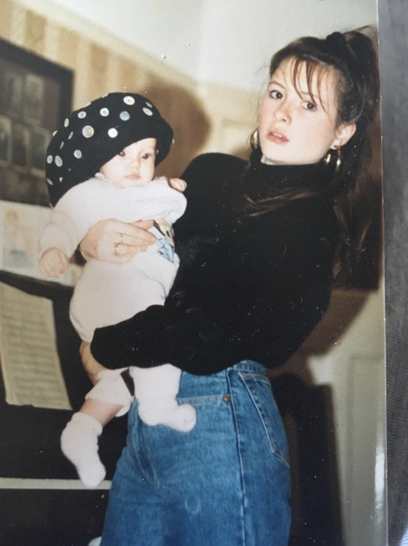 A woman with dark hair wearing a black turtleneck and blue jeans holding a baby dressed in white, with the baby wearing a black hat with white polka dots, in an indoor setting.