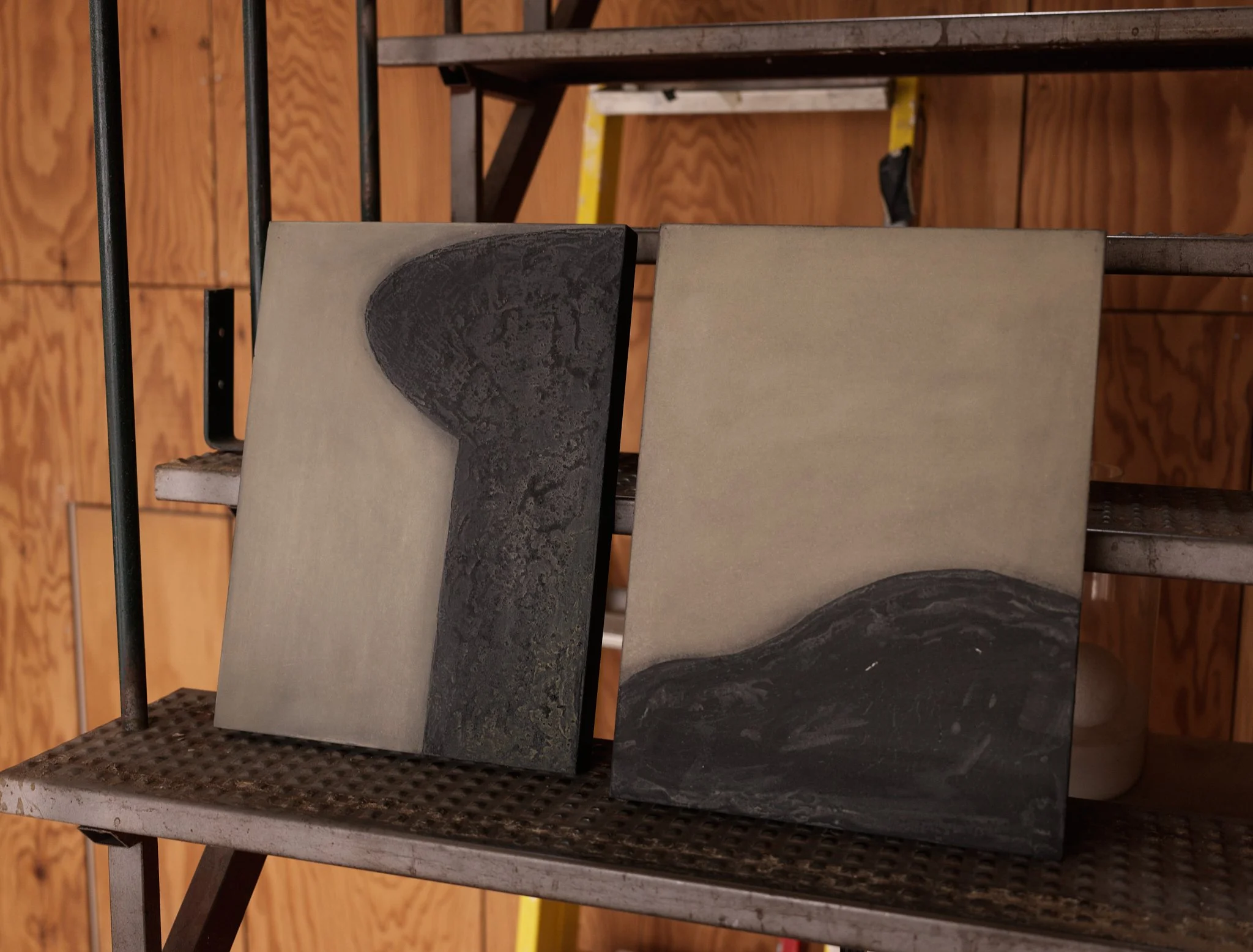 A construction site with wooden walls, metal stairs, and a metal workbench with grid surface, holding three large tiles, one black textured tile, and two beige tiles, in a workshop setting.