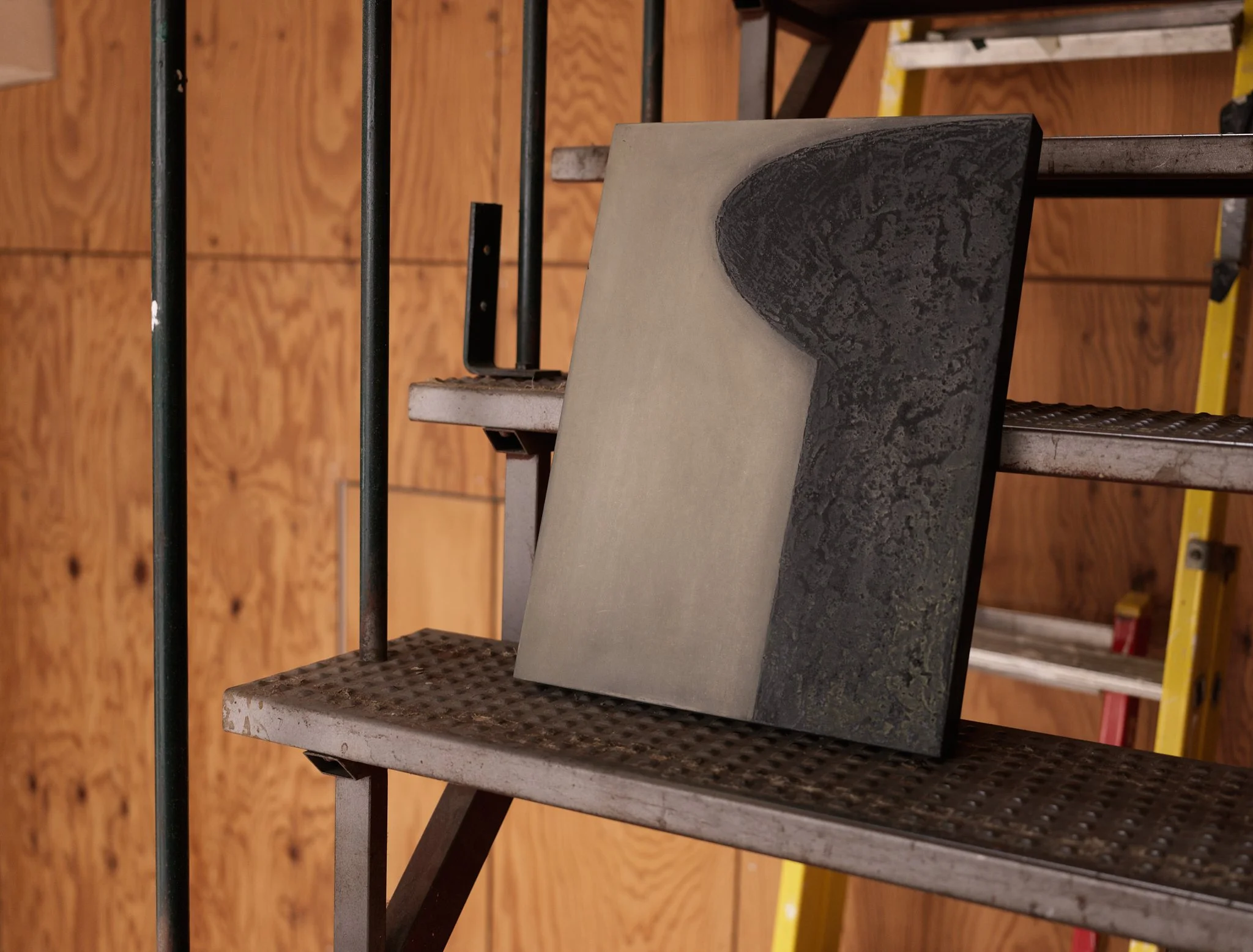 A staircase with metal railings and steps made of black granite and concrete, against a wooden wall and a yellow ladder in the background.