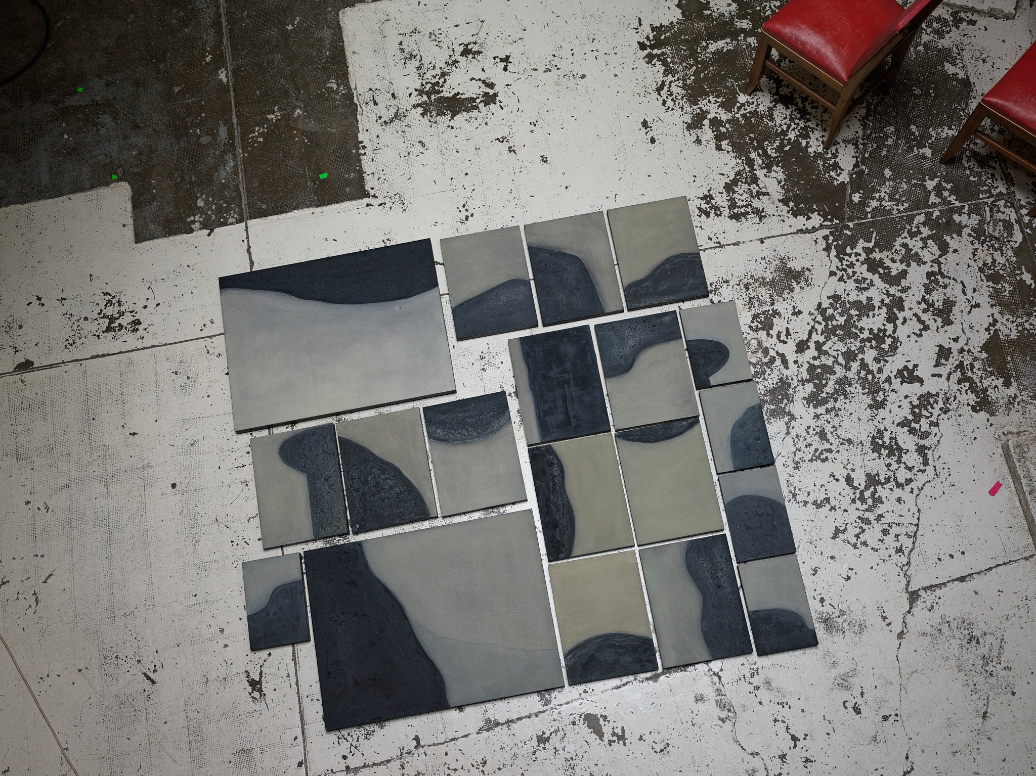 Various square tiles with abstract black and gray patterns are arranged on the floor. Two chairs with red cushions are positioned at the top right corner. The floor appears worn and textured.