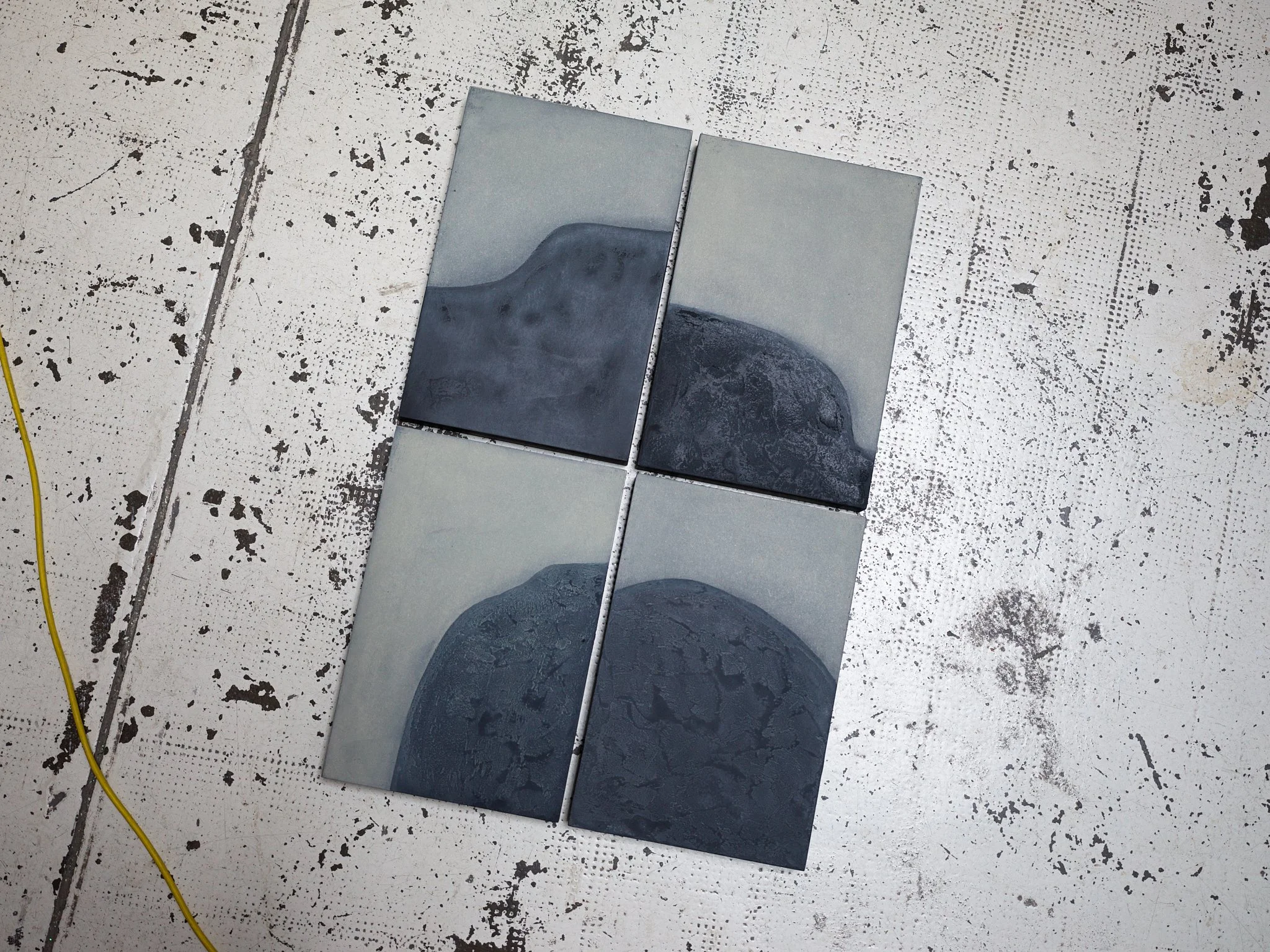 Six tiles, five with gray and black abstract designs and one plain gray, are arranged on a white speckled floor in a room under construction or renovation.