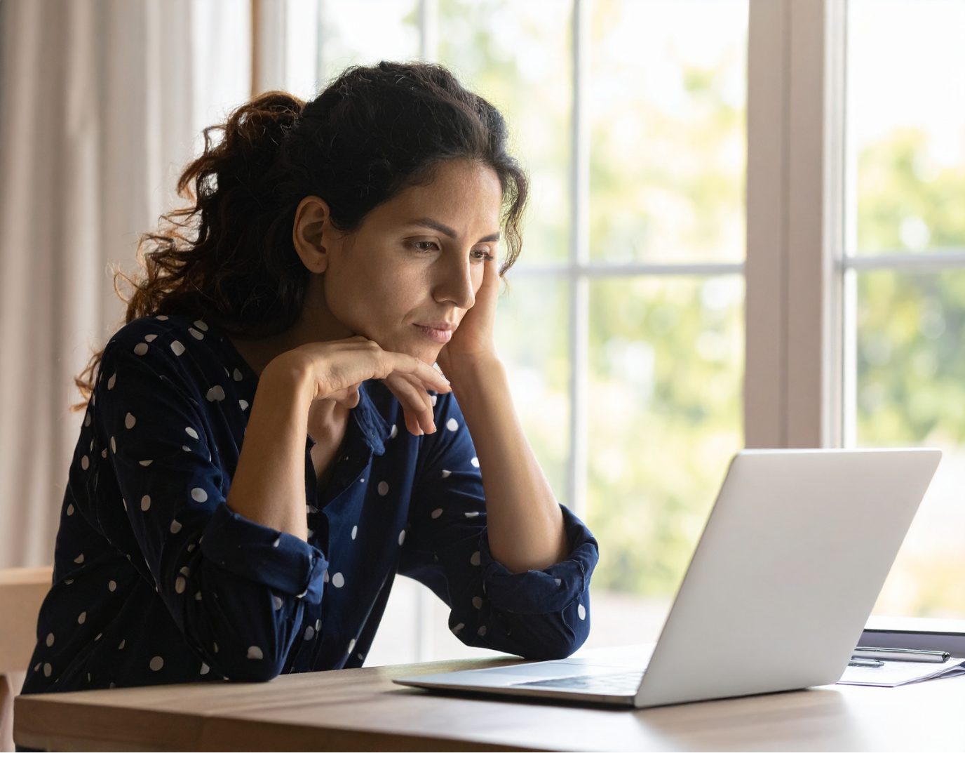 A woman sitting at a desk looking at a laptop with a contemplative expression, near a window with a view of green trees outside.
