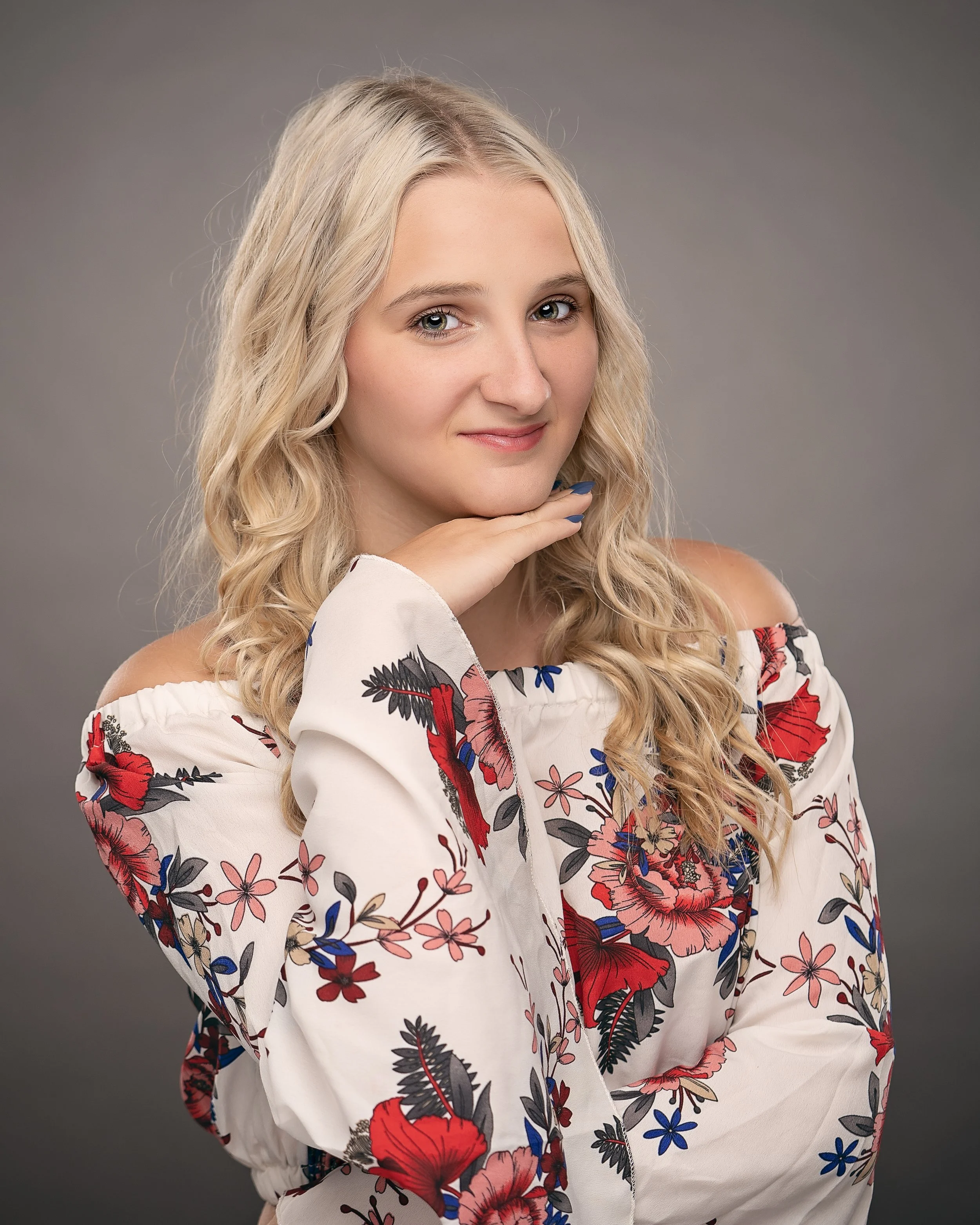 Young woman with long, wavy blonde hair, wearing an off-shoulder white floral blouse, smiling softly against a gray background.