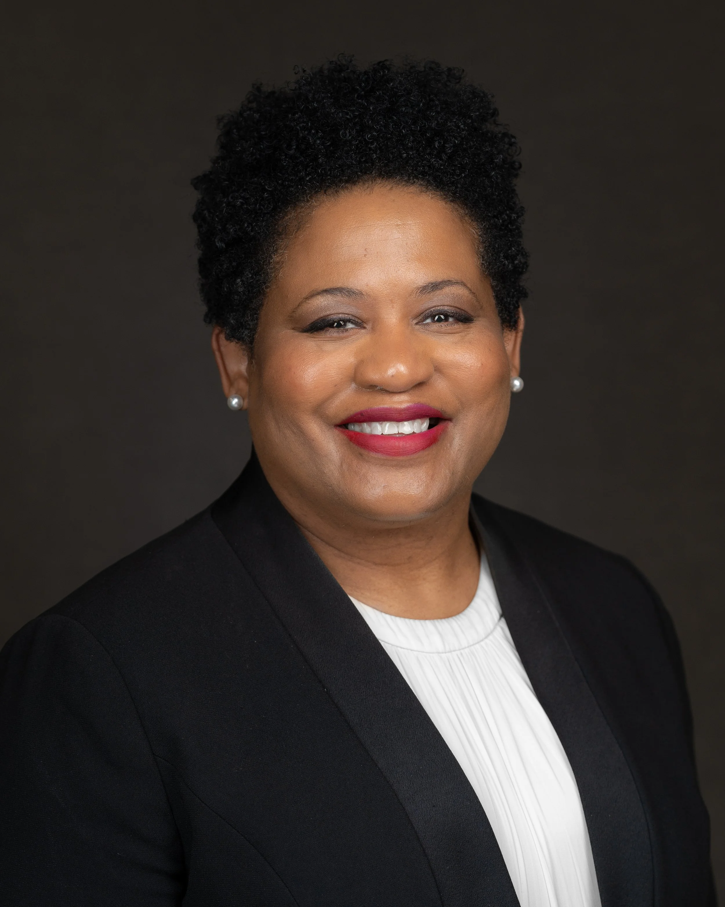 A professional woman with short curly black hair, wearing a black blazer and white blouse, smiling against a dark background.