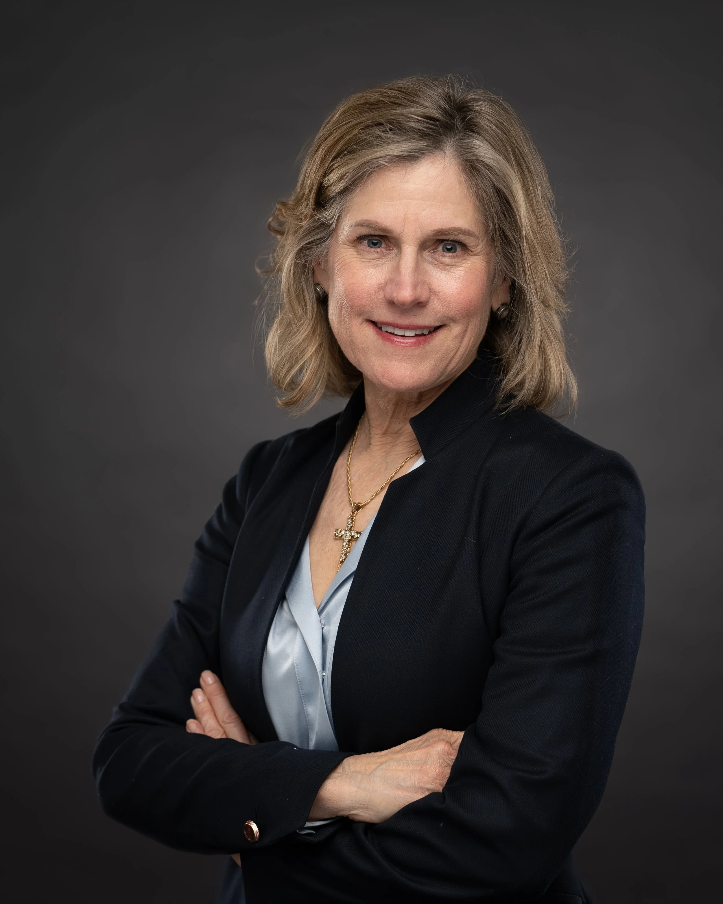 A middle-aged woman with shoulder-length blonde hair, wearing a black blazer, white blouse, and a gold cross necklace, smiling confidently against a dark gray background.