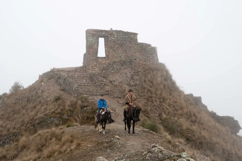 Two people riding horses near the ruins of an ancient stone structure on a hill with foggy weather.
