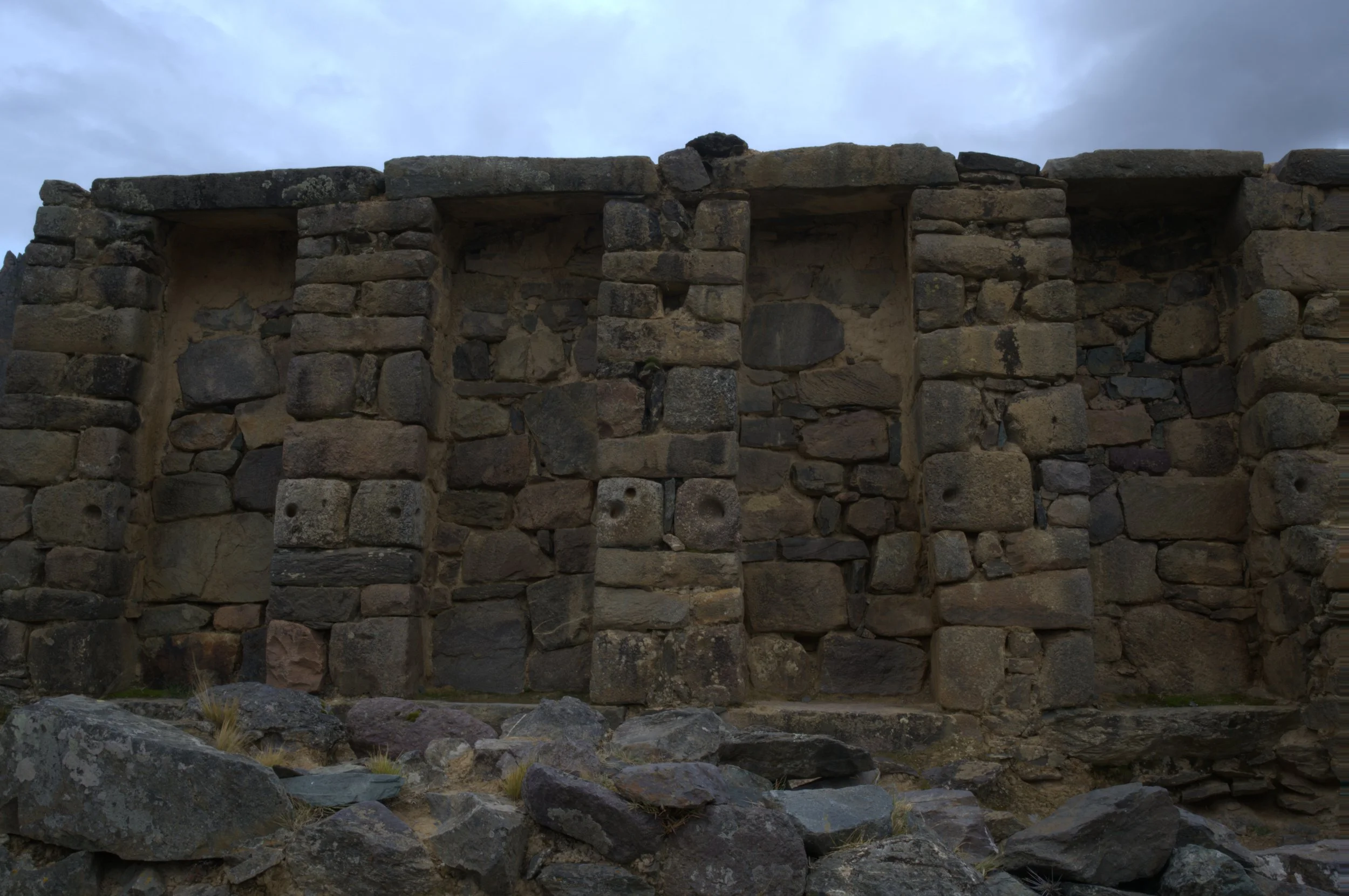 Ancient stone structure with tall, narrow openings, built from irregularly shaped rocks, with rocks scattered in the foreground and a cloudy sky overhead.