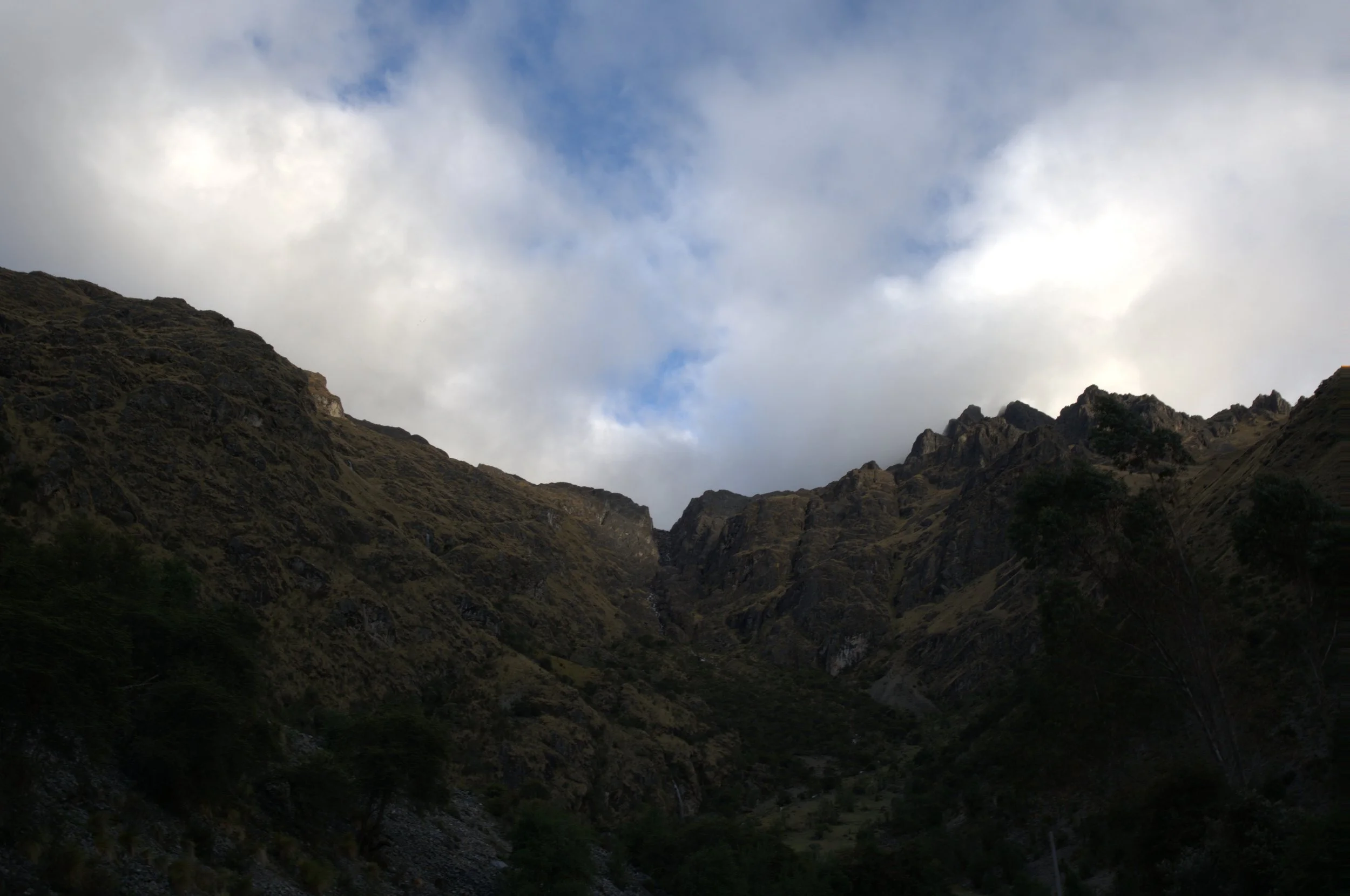 Mountain canyon with steep, rugged slopes covered in sparse vegetation, under a cloudy sky with patches of blue.