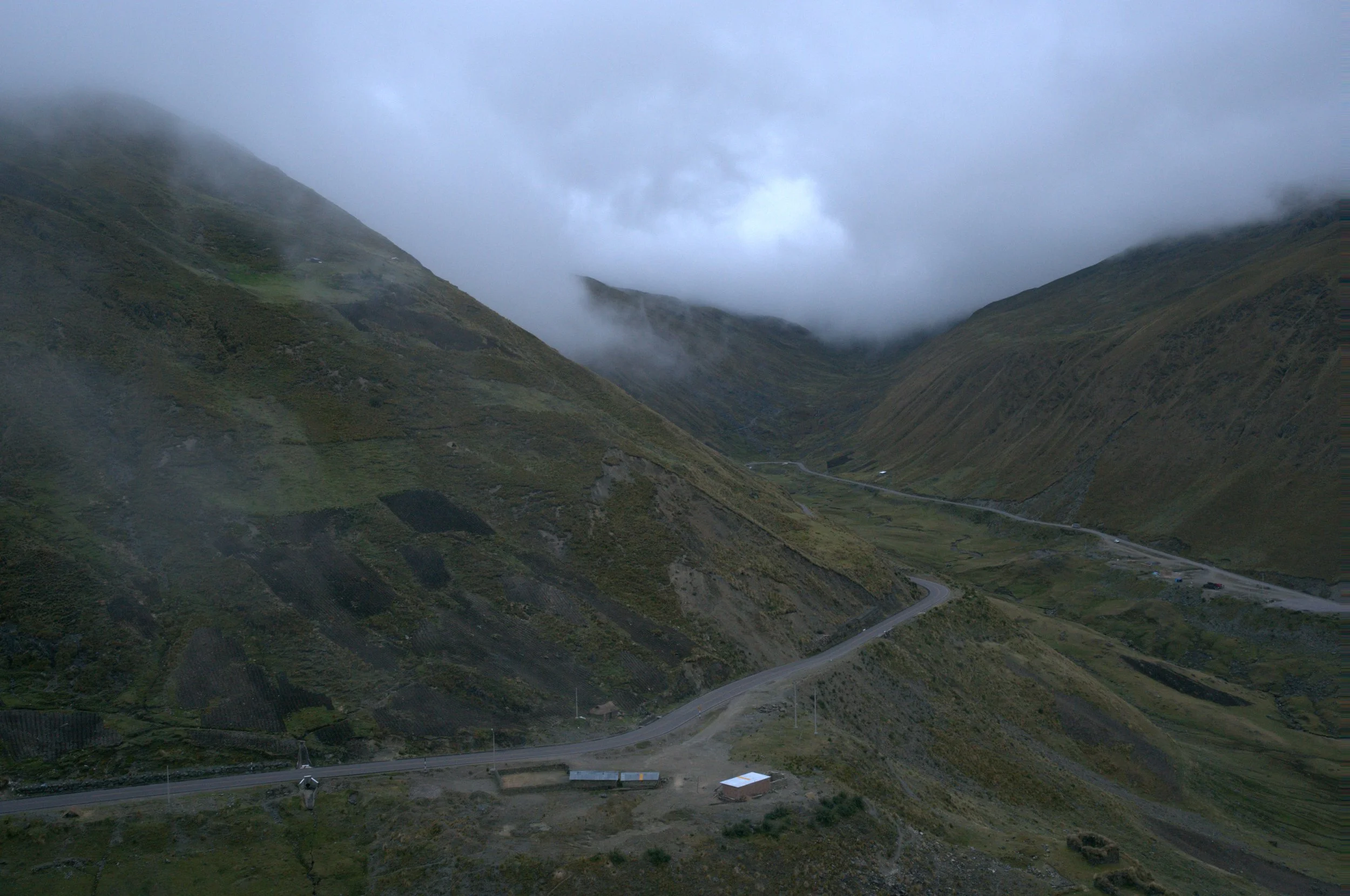 Mountains with fog, winding road, and small buildings at the base, overcast sky.