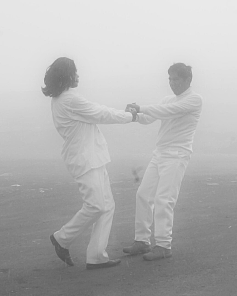 Two people in white outfits holding hands and dancing on a foggy outdoor field.
