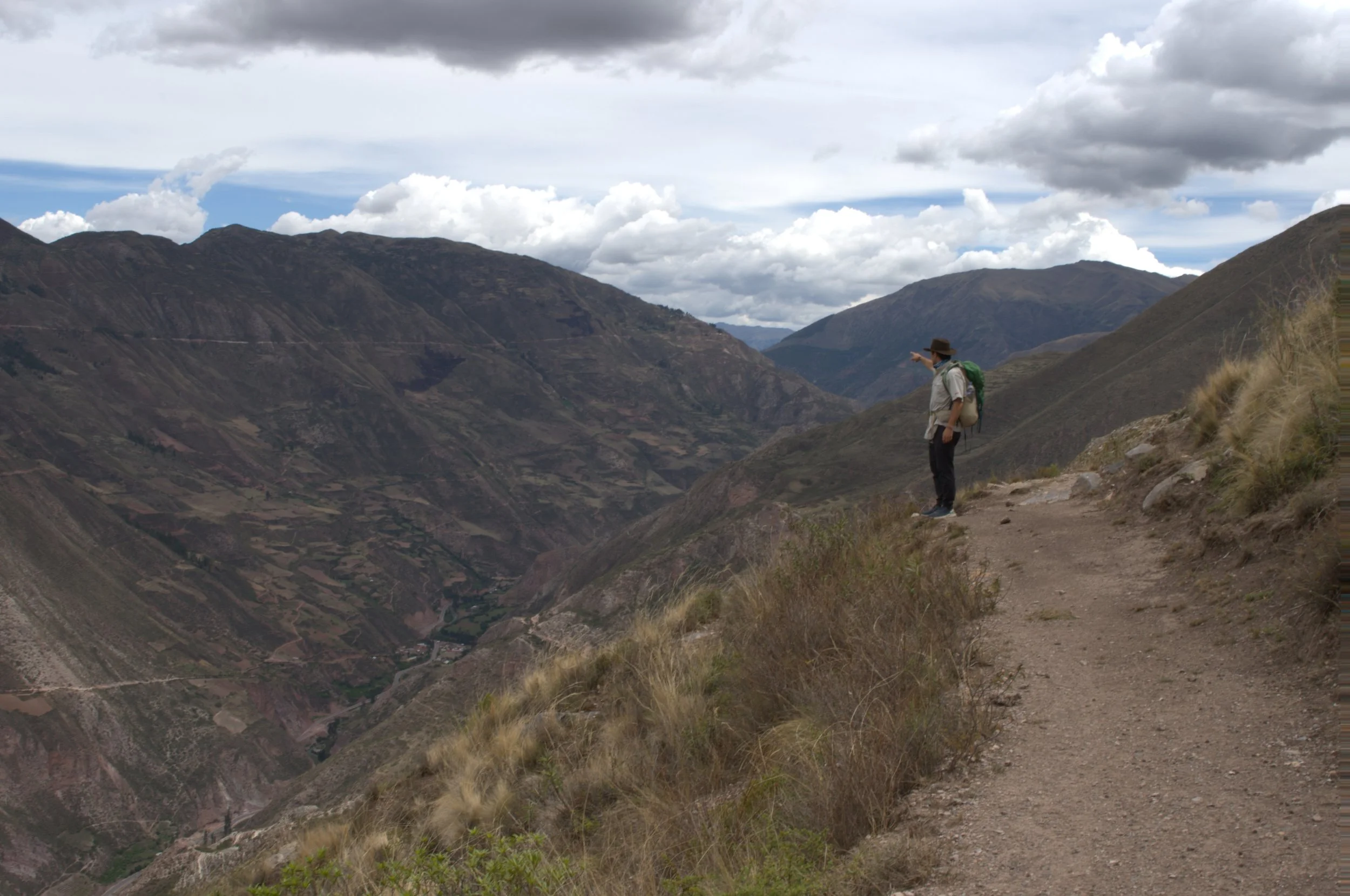 A hiker standing on a trail overlooking a deep valley with mountains in the distance, cloudy sky overhead.