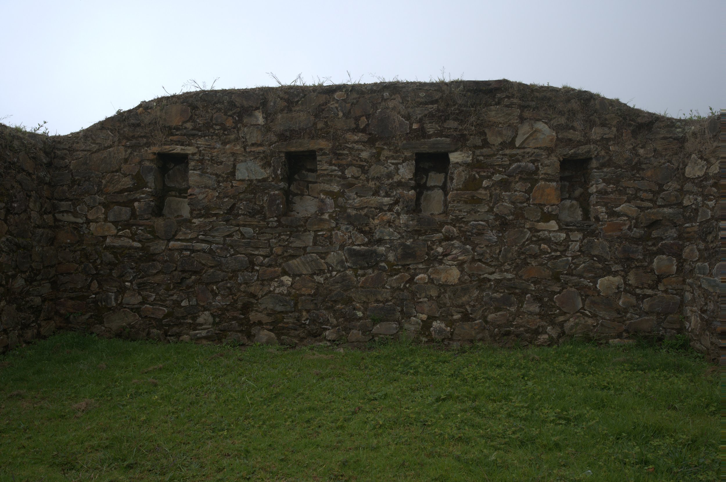 An old stone wall with three small rectangular niches and a grassy ground in front, under a cloudy sky.