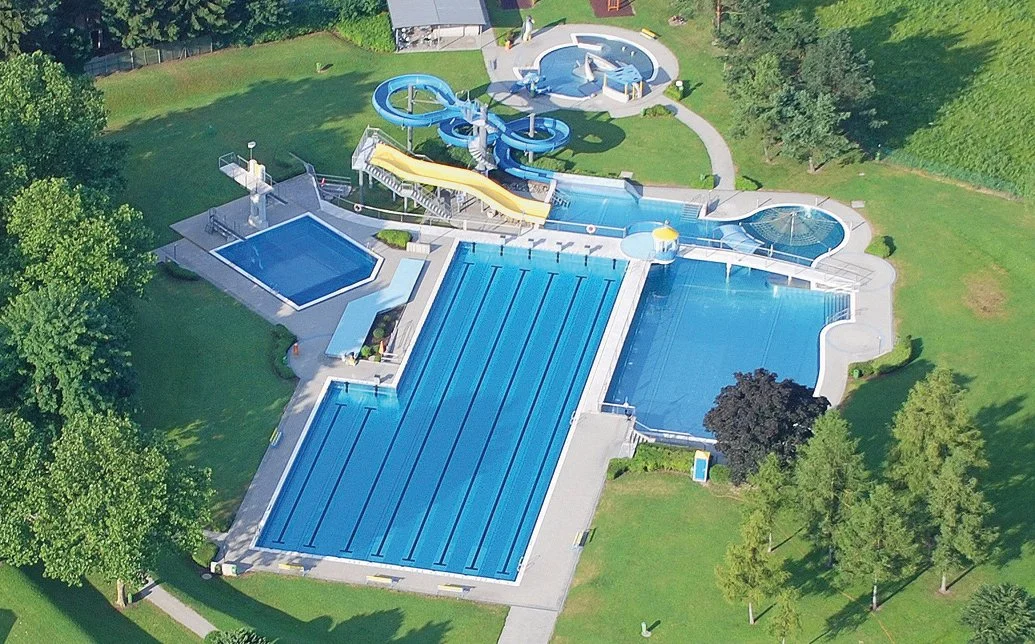 Luftaufnahme eines Wasserparks mit mehreren Schwimmbecken, Wasserrutschen und Wasserstrukturen, umgeben von Grünfläche und Bäumen.