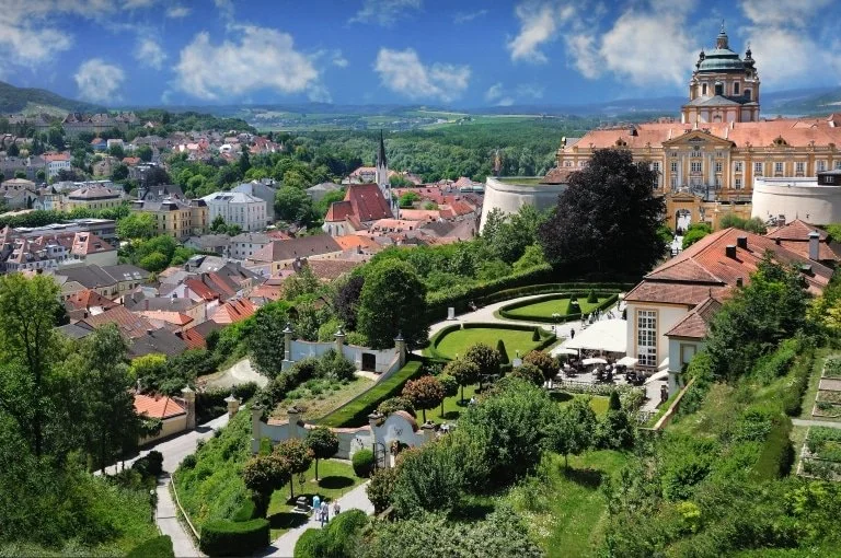 Blick auf eine grüne, blühende Gartenanlage mit gepflegten Hecken und Bäumen, im Hintergrund eine Stadt mit alten Gebäuden, Kirchen und einer großen barocken Palast, umgeben von Hügeln und Wäldern bei schönem Wetter.