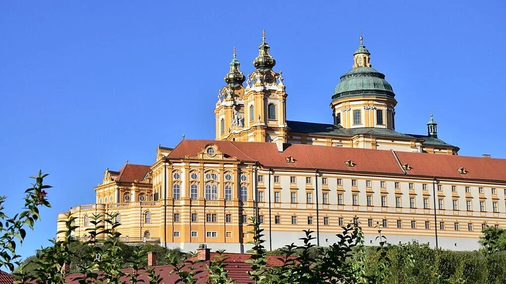 Ein barocker Kirchenbau mit gelb-beiger Fassade, mehreren Türmen und einer großen Kuppel, unter einem klaren blauen Himmel.