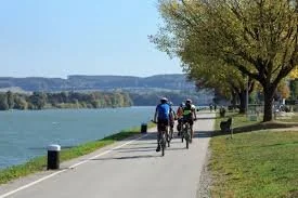 Menschen fahren Fahrrad entlang eines Flussufers mit Bäumen und blauer Himmel im Hintergrund.