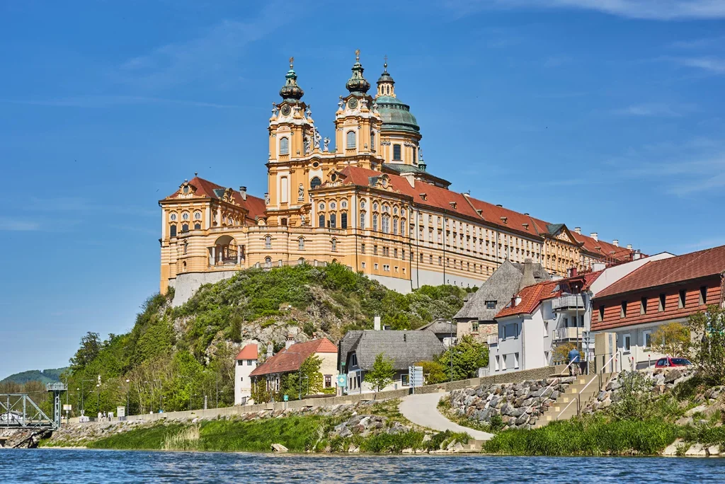 Ein barocker Schloss auf einem Hügel mit Blick auf einen Fluss und umliegende Häuser, unter blauem Himmel.