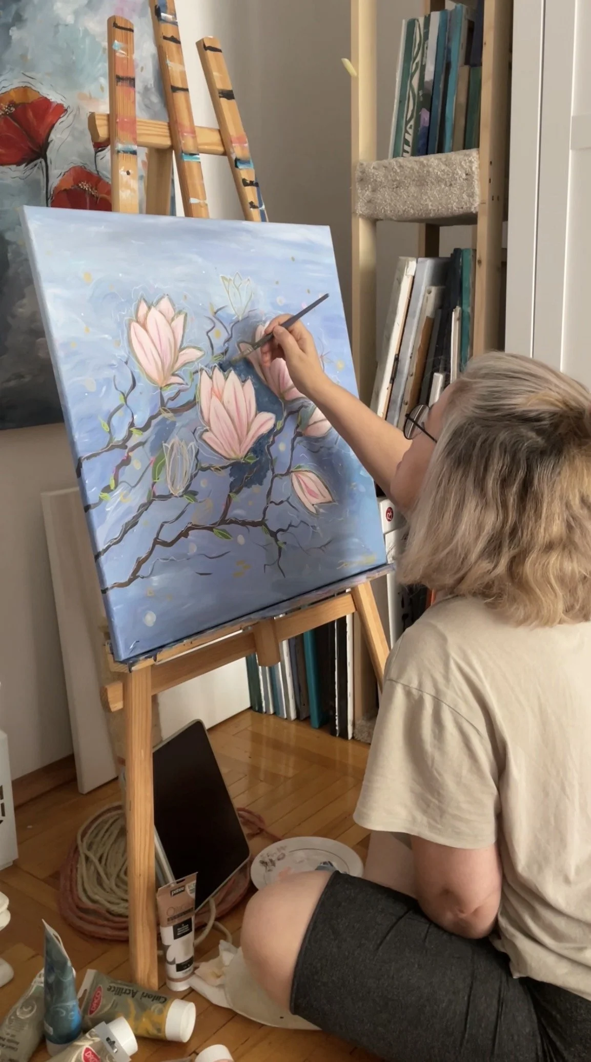 Person painting a floral scene with pink flowers and dark branches on a canvas, sitting on the floor surrounded by painting supplies in an art studio.