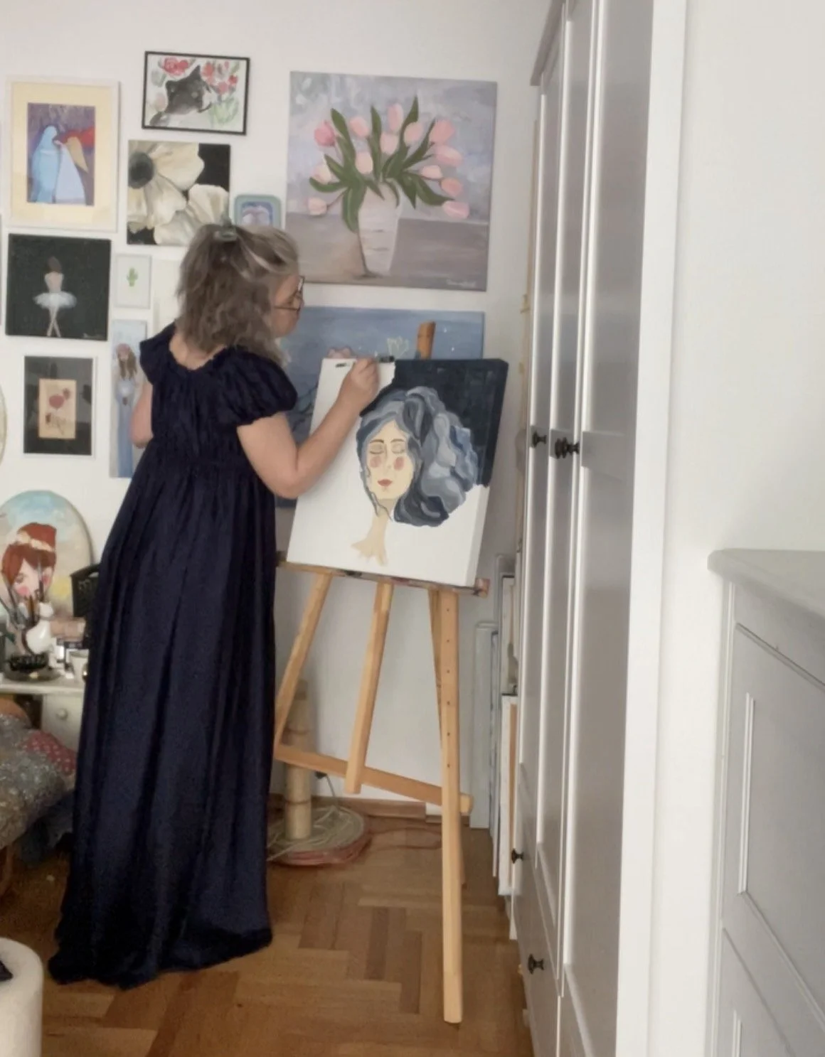 A woman in a long black dress is painting a portrait of a woman with gray and black curly hair on a canvas in a room decorated with various art pieces and paintings on the wall.