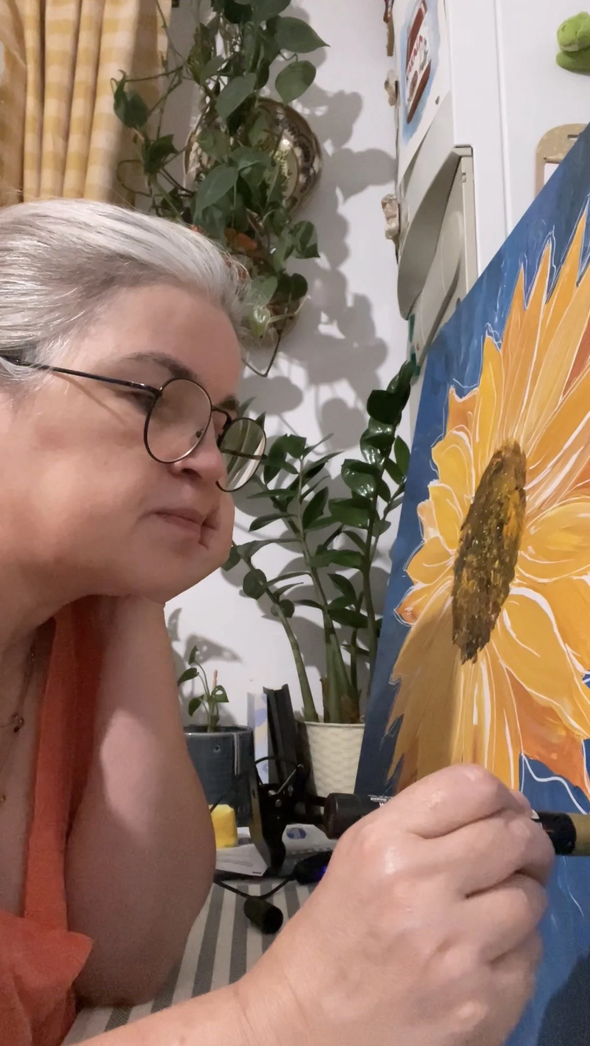 An older woman with glasses and gray hair is painting a sunflower on a canvas.