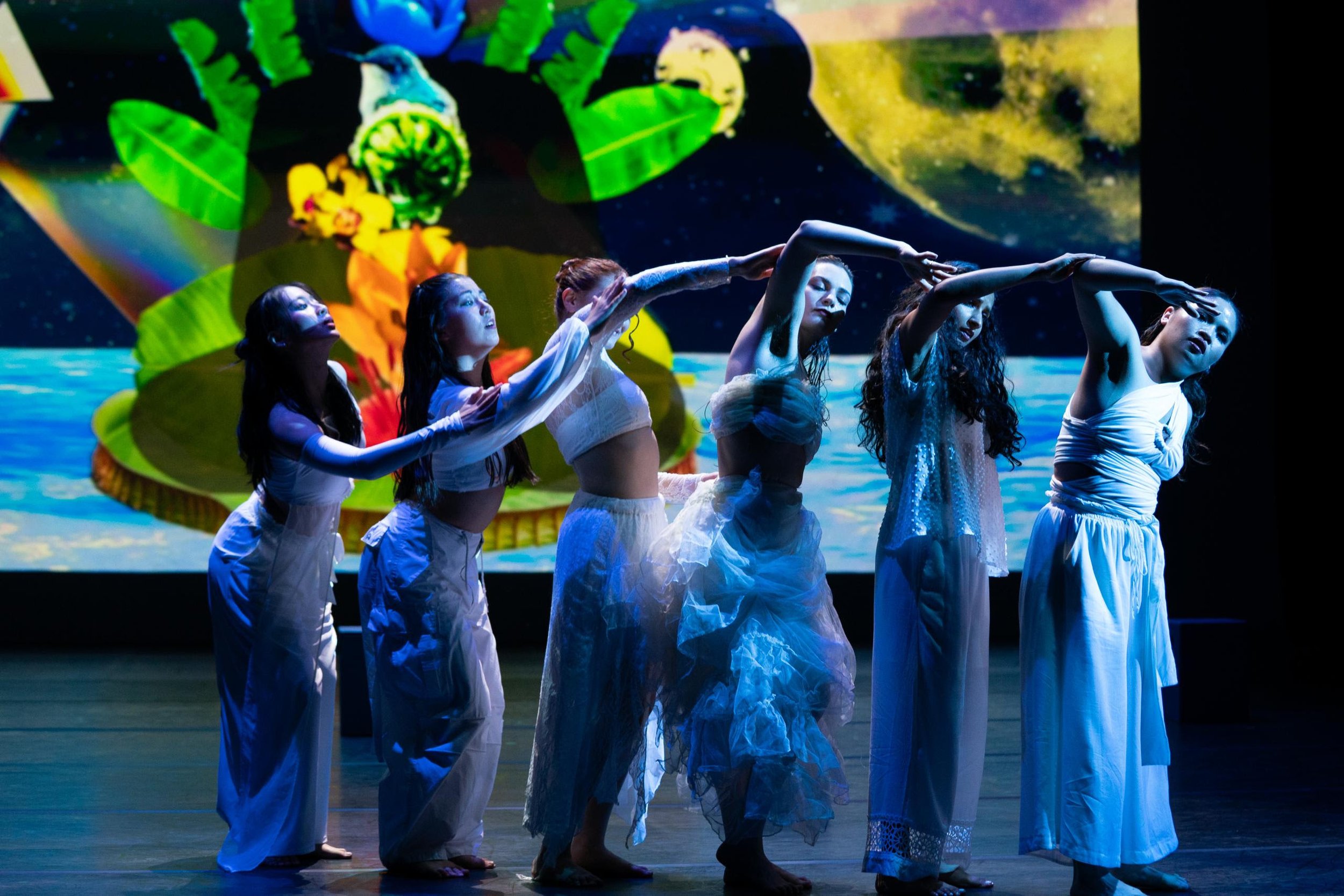 Photograph of six dancers in flowing white outfits standing in a line connecting their arms to make a curving wave. Lit by blue lighting on a stage, with a cosmic digital collage projected behind them. Photo credit: Robbie Sweeny